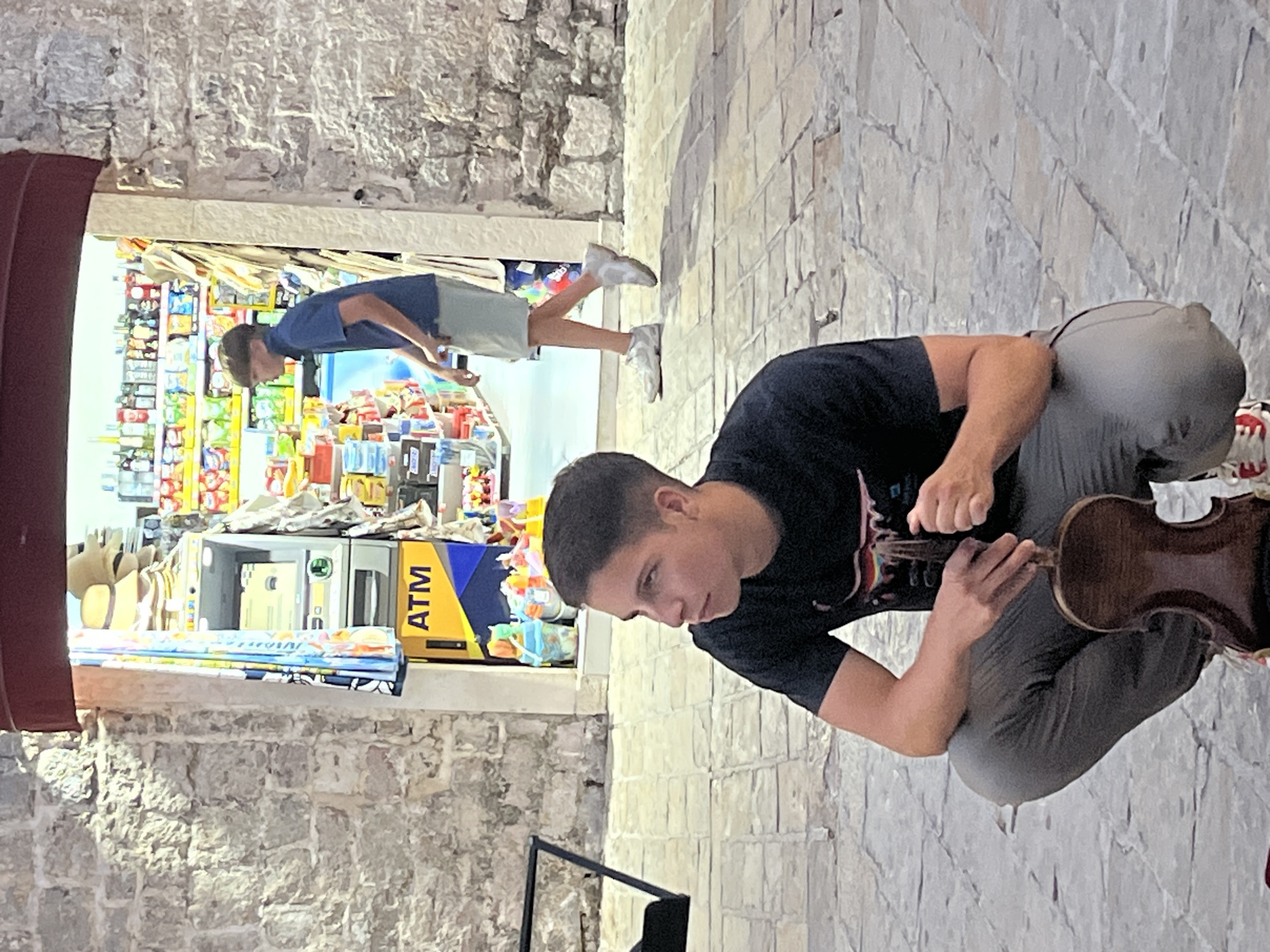 In Budva, Montenegro, an outdoor scene unfolds near a store entrance. In the foreground, a young person with short brown hair is crouched down, holding a violin. They are wearing a black t-shirt and gray pants. To the right, another young person, also with short brown hair, walks away from the store. They are wearing a blue polo shirt and light-colored shorts. The store interior is visible, with shelves stocked with various items and an ATM machine near the entrance. The ground is paved with stone tiles, and the walls are constructed of rough stone blocks.