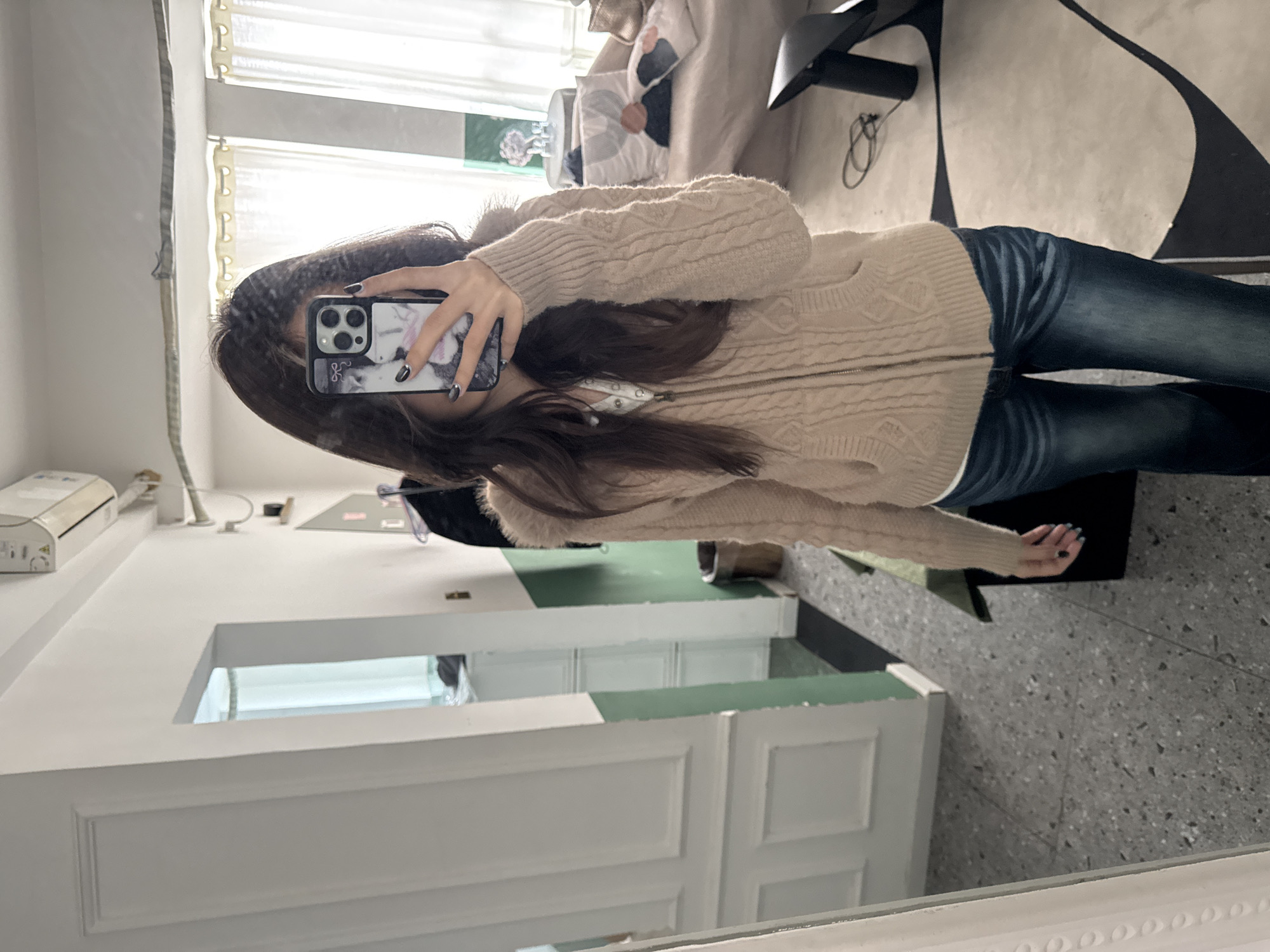 An individual is depicted taking a mirror selfie in an interior domestic setting in Liangjiang, China. The scene focuses on a female subject positioned centrally, reflecting in a large, white-framed mirror.
The subject has long, dark hair and is wearing a light beige cable-knit hooded cardigan over a white top, paired with dark blue denim jeans. Her left hand holds a black smartphone with a multi-lens camera array and a clear case featuring a light-colored graphic design, obscuring her face. Her right hand, with dark-painted fingernails, is visible by her side.
The background consists of an indoor environment. The floor directly in front of the subject is a light gray terrazzo-like material, transitioning to a light-colored rug with dark, curvilinear abstract patterns on the right. Walls are painted in sections: a white wall with panel molding on the left, and a two-toned wall further right with a white upper section and a dark green lower section. A doorway on the left leads into an adjacent room with light-colored furniture and white curtains.
To the right, a light-colored sofa adorned with multiple decorative pillows featuring abstract geometric designs is visible. A small, round, light-colored side table with a clear glass decorative item stands next to the sofa. A dark, angular piece of furniture or an extension of the sofa is partially visible on the far right, with a loose dark cable on the floor nearby. An air conditioning unit is mounted on the upper white wall section, with a small white shelf below it holding an object. A brown cylindrical trash can and a black floor mat are situated against the two-toned wall. A white curtain covering a window on the far right allows natural light into the space. A clothes hanger with dark garments is visible hanging on the wall to the upper right of the subject.