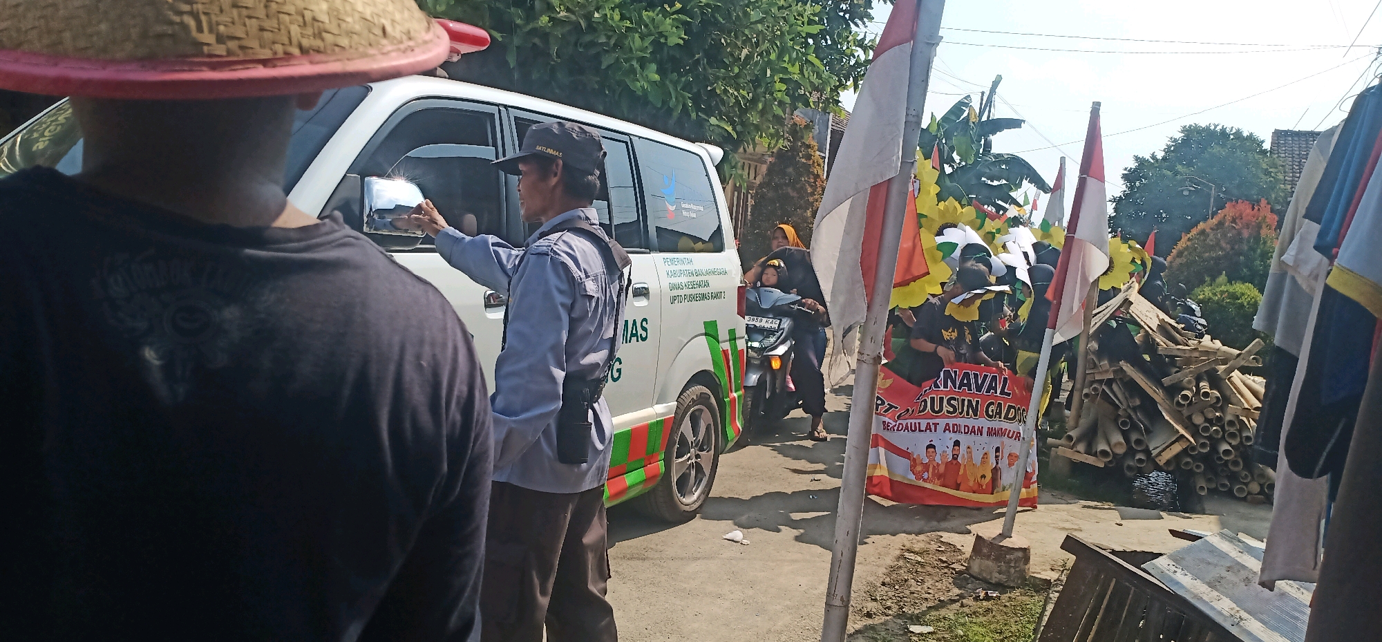 The image depicts a street scene in Kincang, Indonesia, likely during a local event or procession. In the foreground, the back of a person wearing a dark t-shirt and a conical hat is visible, facing a white ambulance or similar vehicle. A man in a light blue uniform and a black cap, identified by the text "SATLINMAS" on his cap, is standing by the driver's side of the vehicle, adjusting the side mirror. The vehicle has text on its side that reads "PEMERINTAH KABUPATEN BANJARNEGARA DINAS KESEHATAN UPTD PUSKESMAS RAKIT 2" and "MAS G". Further to the right, a motorcycle is visible with two people on it, one of whom is holding a child. In the background, flags, decorative elements resembling sunflowers, and a banner are prominent. The banner has Indonesian text that appears to say "KARNAVAL DSUN GADO" and "BERDAULAT ADILAN MAKMUR". A pile of bamboo and wood is stacked to the right of the banner. The environment suggests a sunny day with trees and buildings in the background, indicating a residential or semi-rural area. The overall scene suggests a community gathering or parade.