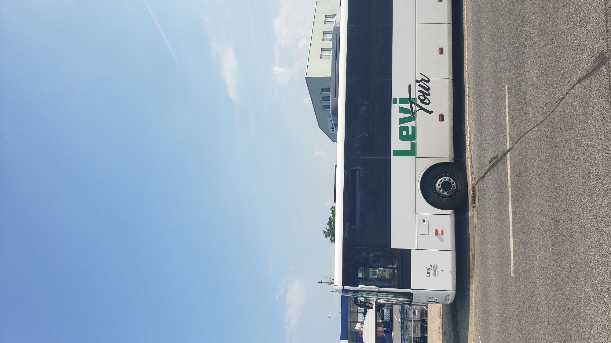 The image shows a white bus with "Levi Tour" written in green on its side. The bus is parked on the side of a road in what appears to be an urban or commercial area. In the background, there is a building with a light green facade and several windows, and what looks like a gas station or a commercial building with a blue canopy. There are also shopping carts visible to the left of the bus. The sky is blue with a few scattered clouds. The overall scene is a daytime outdoor shot. The location is Prešov, Slovakia.