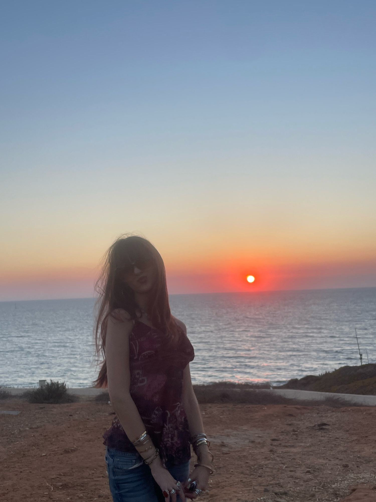 A woman stands on a clifftop overlooking the Mediterranean Sea at sunset. The sky transitions from a clear blue at the top to shades of orange and pink near the horizon. The sun is a bright orange orb low in the sky, partially obscured by the sea. The woman, with long brown hair and sunglasses, wears a patterned tank top and jeans. She is adorned with several bracelets and rings. The ground in the foreground is covered with reddish-brown soil and sparse vegetation. The sea is calm with gentle waves. The location is the Herzliya beach area, Israel, known for its picturesque sunsets.