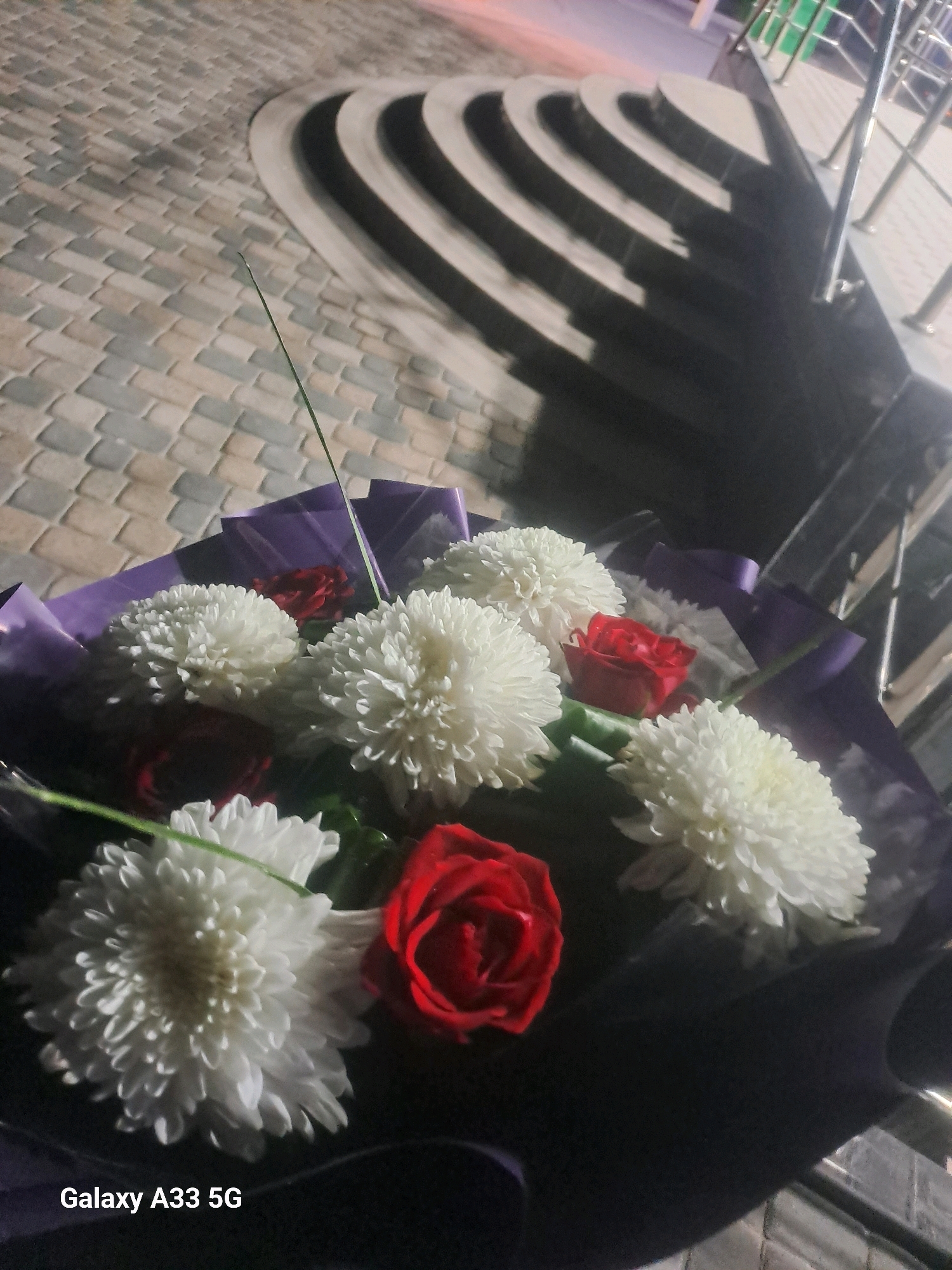 A bouquet of white chrysanthemums and red roses is arranged with purple wrapping. The bouquet is placed on a dark surface, possibly a table or ground, in front of a curved staircase with dark steps and a lighter tiled surface. The scene appears to be outdoors, with some lighting from the side casting shadows. The lower left corner of the image displays the text "Galaxy A33 5G".