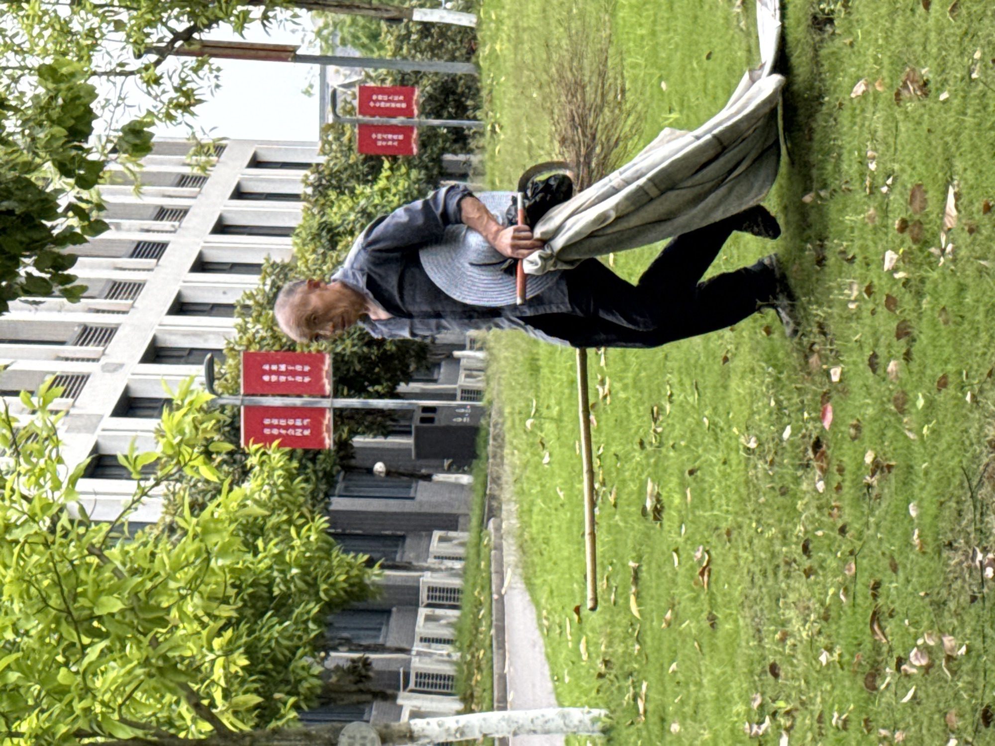 An elderly man is walking on a grassy lawn in what appears to be an outdoor area. He is wearing dark pants, a blue and grey long-sleeved shirt, and black shoes. He holds a wide-brimmed straw hat and a light-colored cloth or shawl. He is also carrying what appears to be a thin wooden pole. Behind him is a walkway, bordered by green trees and foliage and a modern building with multiple windows. Red signage with writing is visible between the building and the trees. The location may be Jiangjin, China.