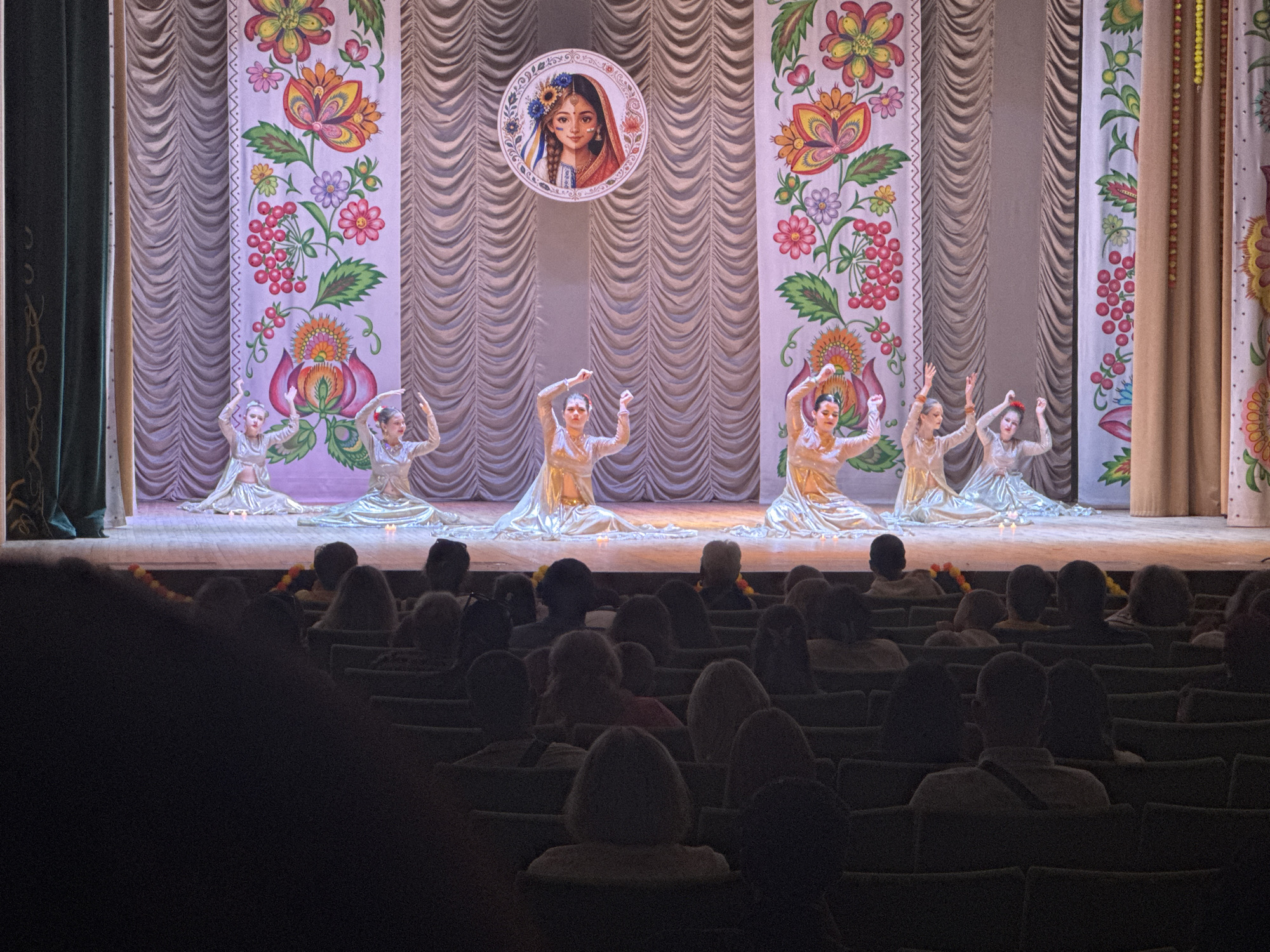 The image depicts a stage performance viewed from an audience perspective. On stage, five young women in matching golden, shimmering costumes are performing a dance. They are positioned in a semi-circle, with their arms raised and their bodies angled in graceful poses. The stage backdrop features ornate, colorful floral banners with a muted, textured curtain behind them. Centered at the top of the backdrop is a circular image of a young woman with traditional attire and braided hair. The foreground is dominated by the silhouettes of the audience, seated in rows of dark chairs, suggesting the performance is taking place in a theater or auditorium. The lighting on stage is bright, illuminating the performers and the backdrop, while the audience area is dim. The overall scene captures a moment of artistic performance and audience engagement.