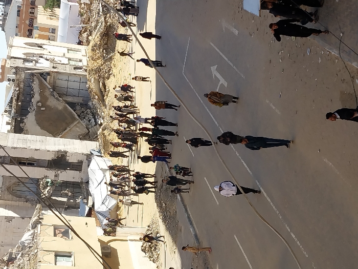 The image displays an urban street scene in Khan Yunis, Palestinian Territory, characterized by significant structural damage. In the background, a large multi-story building on the right exhibits extensive collapse, with a substantial portion of its facade missing, revealing interior structures and a mound of rubble at its base. An adjacent building to the left also shows damage to its upper sections. Extensive debris and rubble piles are scattered across the background, among which a large white tent structure is visible. The foreground features a paved street with white lane markings, including a directional arrow indicating a right turn and a straight-ahead path. A thick, light-colored hose or cable runs diagonally across the street. Numerous individuals, including adults and children, are present. Many are walking on the street, while others are gathered in groups among the rubble and damaged structures in the mid-ground. Their attire consists primarily of casual clothing, including jackets and hoodies. Overhead wires are visible across the upper portion of the frame under a clear sky.