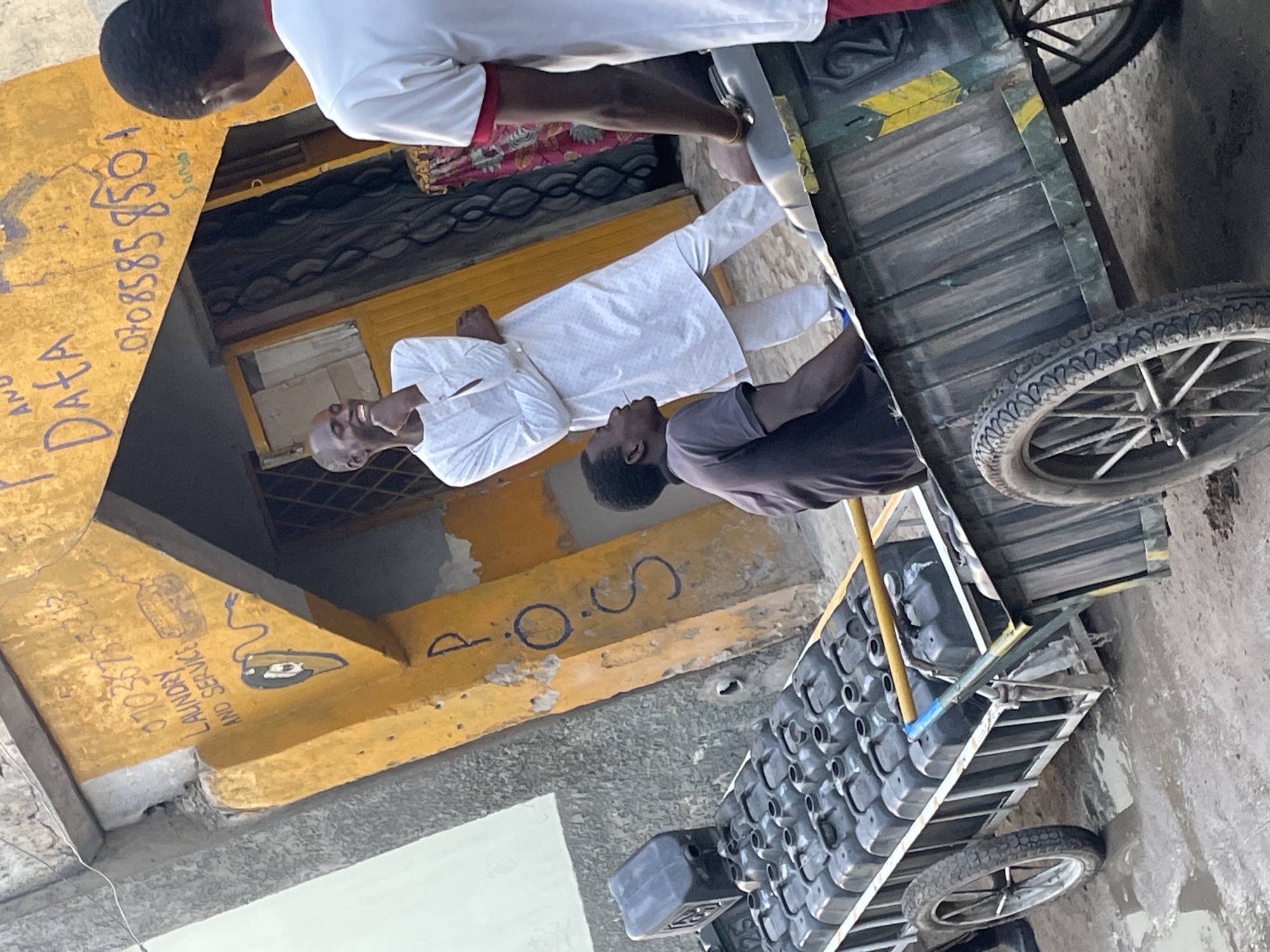 The image captures a scene in what appears to be a city street, featuring a building with yellow-painted walls and visible signage. A man in white clothing stands near the building, interacting with a person in a dark shirt. Above them, a partial view of another individual in a white shirt with red trim is visible. A wooden cart on wheels, loaded with a large number of identical black containers, occupies a significant portion of the frame. The cart is situated close to the people and building. Visible on the yellow wall are painted numbers "07085858501" and text related to "LAUNDRY AND SERVICE 0103075". The environment seems to be a typical urban setting with a mix of architectural styles. The image's location is Unknown, Unknown.