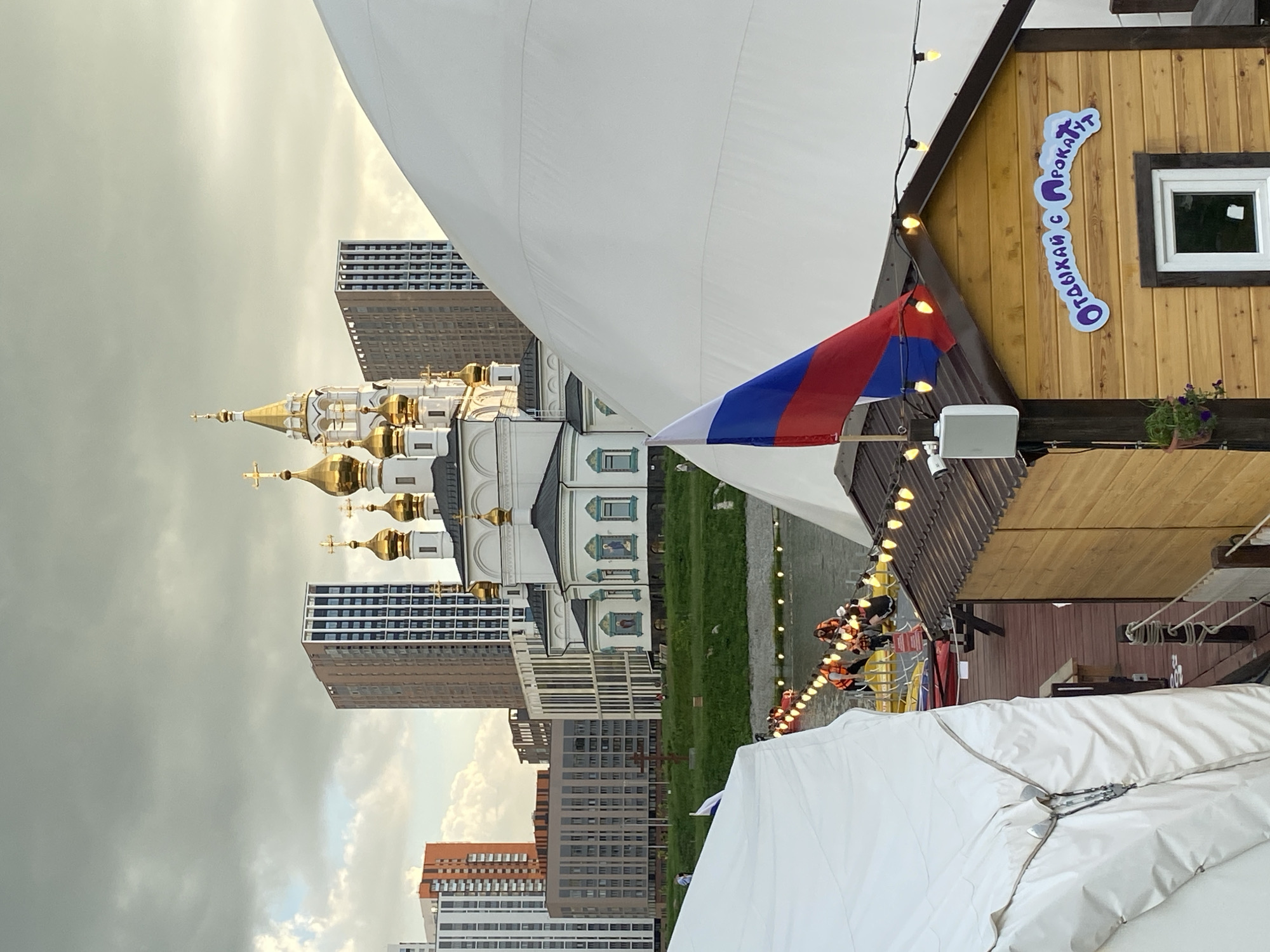 In Yekaterinburg, Russia, on a cloudy day, a view overlooks a waterfront area featuring a white domed structure and a cluster of modern high-rise buildings in the background. A prominent Orthodox church with golden domes stands in the mid-ground. In the foreground, a small wooden structure with a brown roof and string lights is visible, bearing a sign with Russian text that translates to "Relax with a Ride." A Russian flag is positioned on a pole attached to this structure. A group of people wearing orange life vests are on a yellow raft in the water, suggesting a recreational activity. White tents are visible in the left and right foreground, partially obscuring the scene.