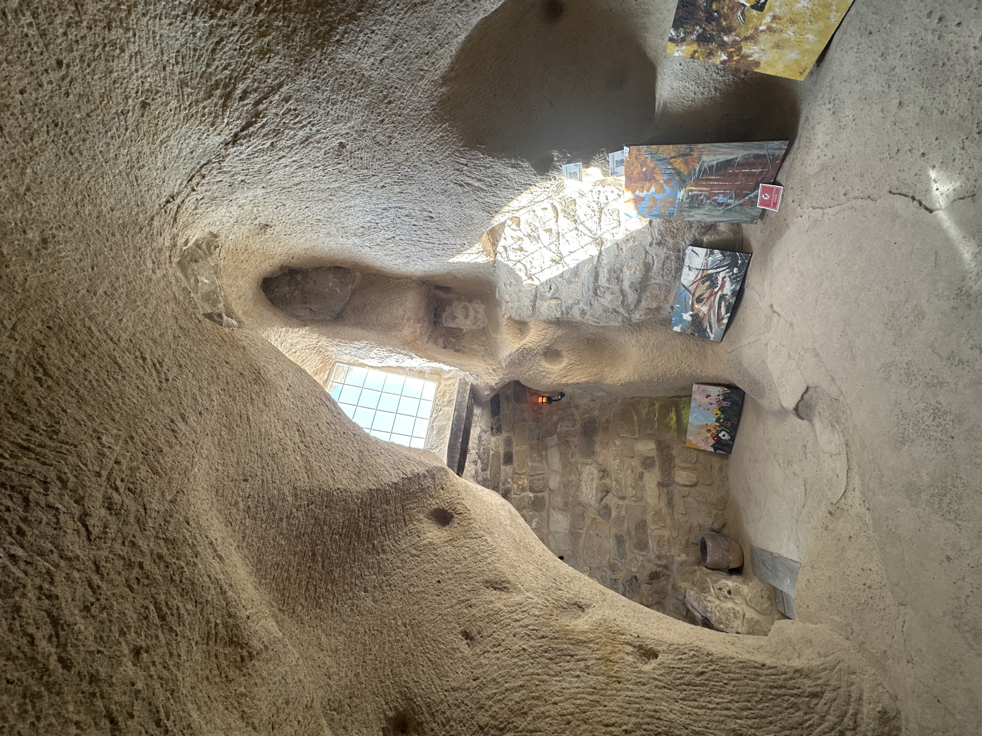 The image depicts the interior of a cave dwelling, likely a former residence or a natural formation, in Uçhisar Beldesi, Turkey. The rough, textured walls suggest that the space has been carved or sculpted from the natural rock. Natural light streams in from a multi-paned window located high up in the cave's ceiling, illuminating the earthy tones of the interior.
The cave floor is uneven and appears to be made of packed earth or stone. Several paintings are displayed within the space, leaning against the rough-hewn walls and on the floor. One painting, towards the back left, is smaller and features a colorful depiction of flowers against a dark background. Further to the right, larger canvases showcase abstract or landscape scenes with a focus on warm colors. A small, dark-colored pot or vase rests on a ledge near the back wall. A simple, rustic light fixture hangs from the stone wall near the window. The overall impression is one of a rustic, arts-focused dwelling within a natural cave environment.