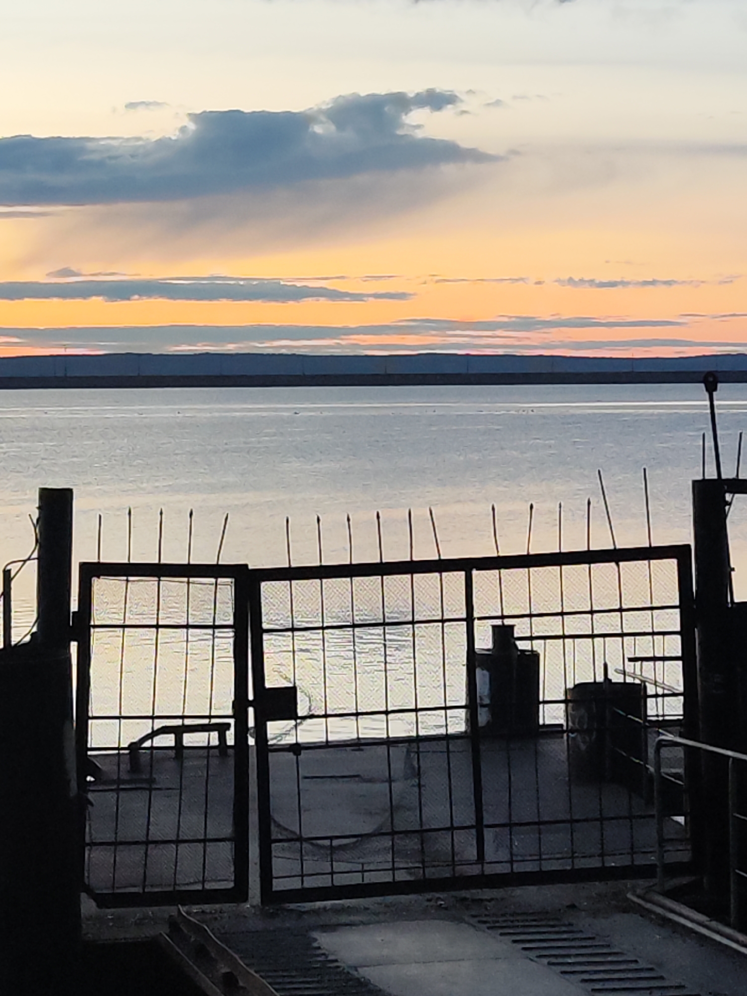 The image shows a view of the Kama River from the ferry terminal in Chaikovsky, Russia. A dark, wrought-iron gate is in the foreground. The river occupies the middle ground with a distant shoreline. The sky is visible above, displaying clouds and a sunset with bands of orange and gray hues. The gate is partially open, revealing a portion of the ferry deck, including a bench and some equipment. The general tone is subdued and quiet.