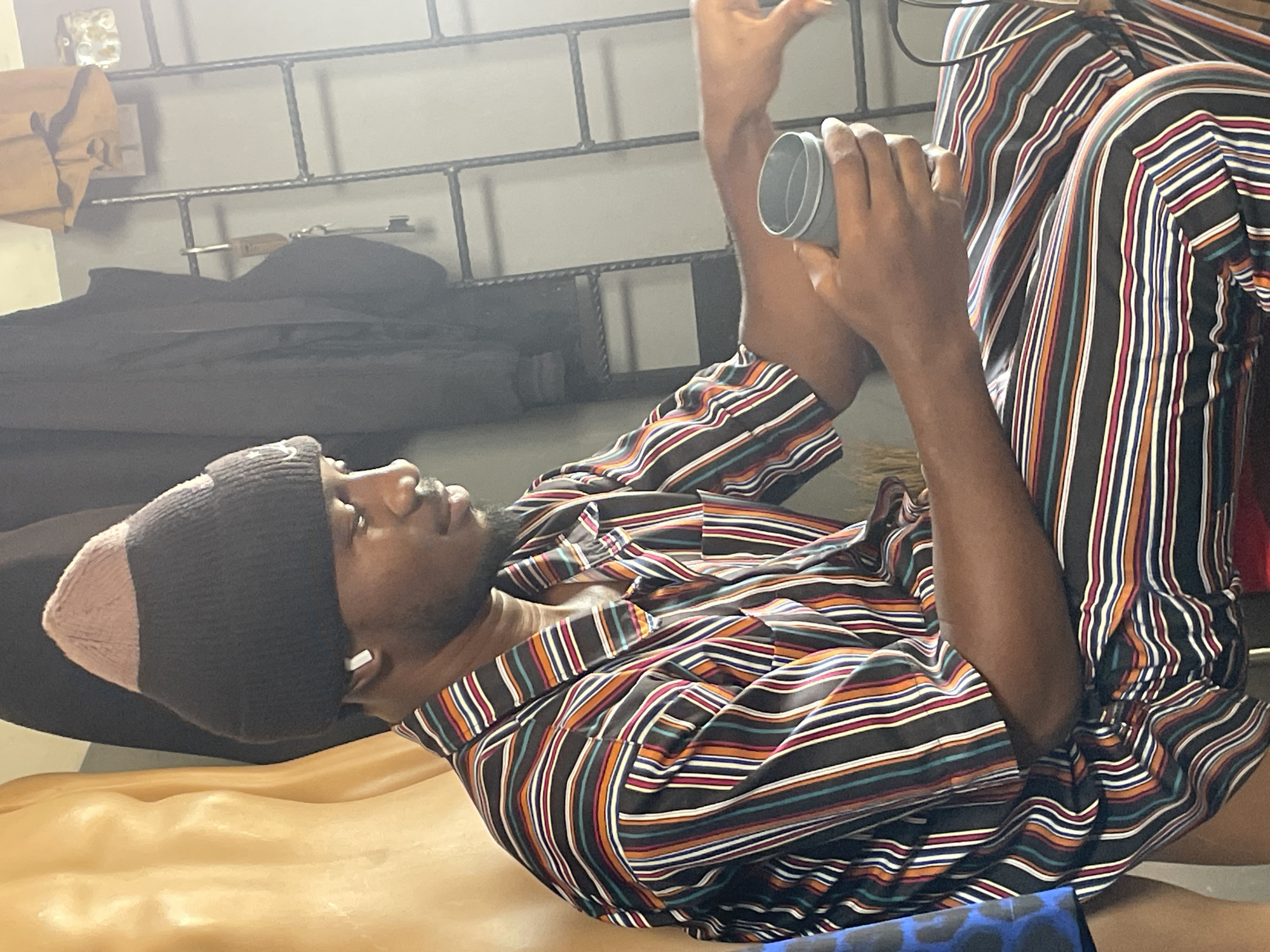 The image shows a person wearing a striped shirt and pants, with a beanie on their head. They are holding a grey object in their hands and appear to be looking at it. The background includes a metal rack and some dark clothing. The lighting suggests it is indoors, possibly daytime. There is no visible text in the image. The setting appears to be a room in Bukuru, Nigeria. The person's posture and facial expression are difficult to discern clearly due to the angle and resolution of the image.