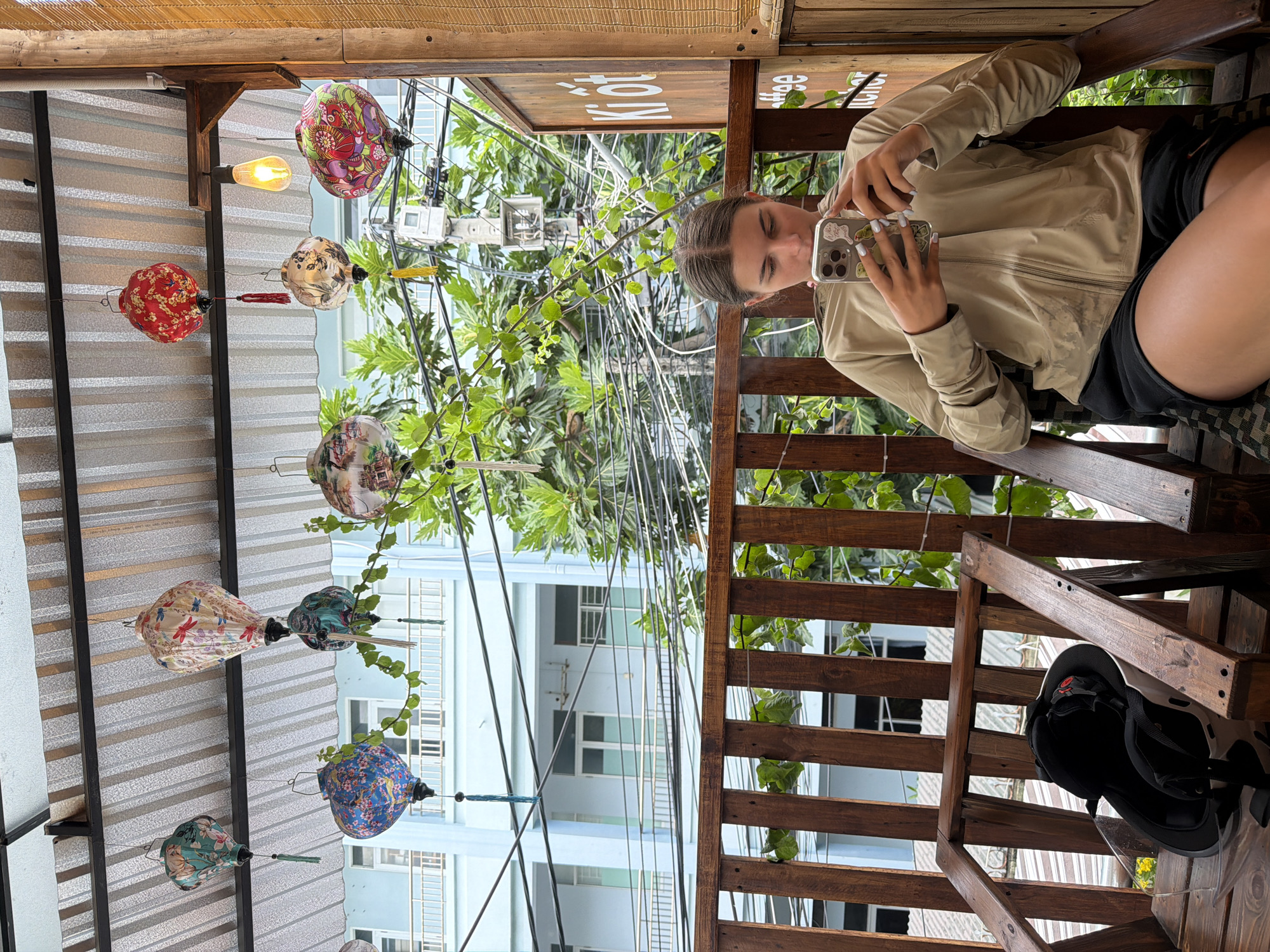 A young woman with brown hair is sitting on a wooden chair, looking at her phone. She is wearing a tan zip-up jacket and black shorts. The chair is on a wooden balcony with a wooden railing. In the background, there are several colorful lanterns hanging from the ceiling. Beyond the balcony, there is a light blue building with windows and balconies, as well as lush green trees and power lines. A motorcycle helmet is on the seat next to the woman. The scene appears to be outdoors in a city, likely in Nha Trang, Vietnam, given the lanterns and the architecture.