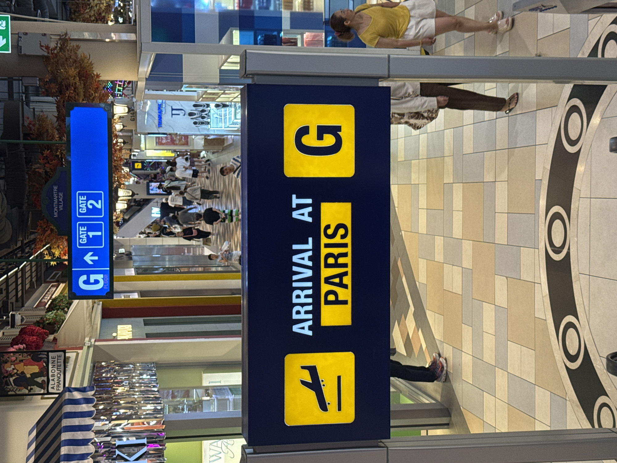 The image captures a scene within a commercial establishment, likely a shopping mall or airport terminal, in Pattaya City, Thailand. A prominent blue sign with white lettering indicates "MONTMARTE VILLAGE" and displays directional arrows pointing to "GATE 1" and "GATE 2". Below this, a larger dark blue sign features a yellow square with an airplane icon, and the text "ARRIVAL AT PARIS" in white and yellow lettering. To the right of this text, another yellow square contains the letter "G" in large black font.
Several individuals are visible in the background, some walking and others standing. On the right side of the image, a woman wearing a yellow top and white shorts is standing. On the left, a person in dark pants and red and white shoes is partially visible. The flooring is a mosaic of beige, grey, and cream square tiles, with a decorative circular pattern in black and white in the foreground. The environment appears to be indoors, with artificial lighting and shops or displays lining the walkways. The overall atmosphere suggests a public space with ongoing activity.