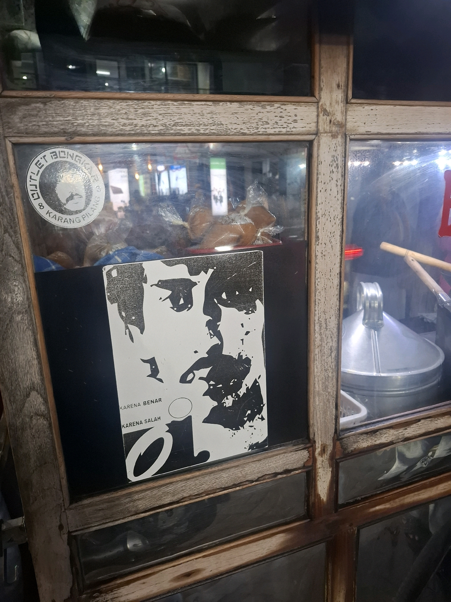 The image shows a close-up of a wooden display case with glass panes. A black and white poster featuring a stylized portrait and text in Indonesian is affixed to the inside of one of the glass panes. Text on the poster reads "KARENA BENAR" and "KARENA SALAH". A circular sticker with text "OUTLET BONGKAR & KARANG PILANG" and a depiction of a man's face is on another pane. The background through the glass shows blurred lights and objects, suggesting an indoor setting or a shop. There are no people visible in the image, and no specific location in Perumahan Puri Indah, Indonesia can be definitively identified from the visual information.