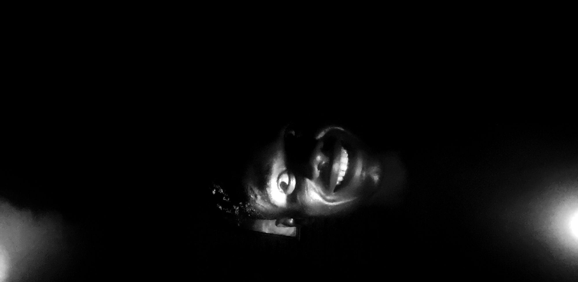 A low-angle, black and white photograph depicts a person's face illuminated against a dark background. The person is looking upwards and smiling widely, revealing their teeth. The image appears to have been taken in low-light conditions. There is no indication from the image that this scene is located in Narayi, Nigeria.