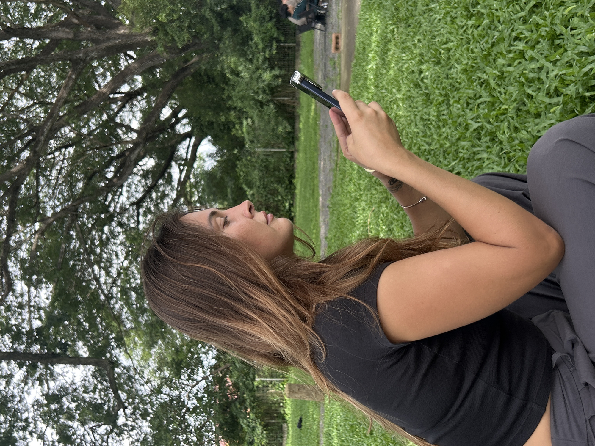 In Tambon San Phi Suea, Thailand, a young woman with long, wavy brown hair is captured from the side, looking down at her smartphone. She is wearing a black t-shirt and dark gray pants, seated outdoors on a patch of vibrant green grass. Her left arm rests on her lap, and her right hand holds the phone. Behind her, a large tree with an intricate canopy of branches and leaves dominates the background. In the distance, a blurred scene shows two people sitting at a table with chairs. The environment suggests a park or outdoor recreational area.
