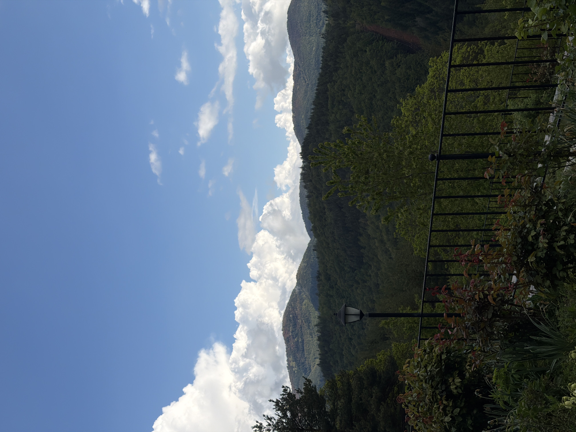 A slightly rotated outdoor shot depicts a mountainous landscape under a partly cloudy, blue sky. The mountain range, heavily forested, is interspersed with patches of exposed rock and earth. A black metal fence runs parallel to the mountains and is partially obscured by greenery and foliage. A black lamppost is also visible in the foreground. The location is implied to be near or within Кирково, Bulgaria. No people are visible.