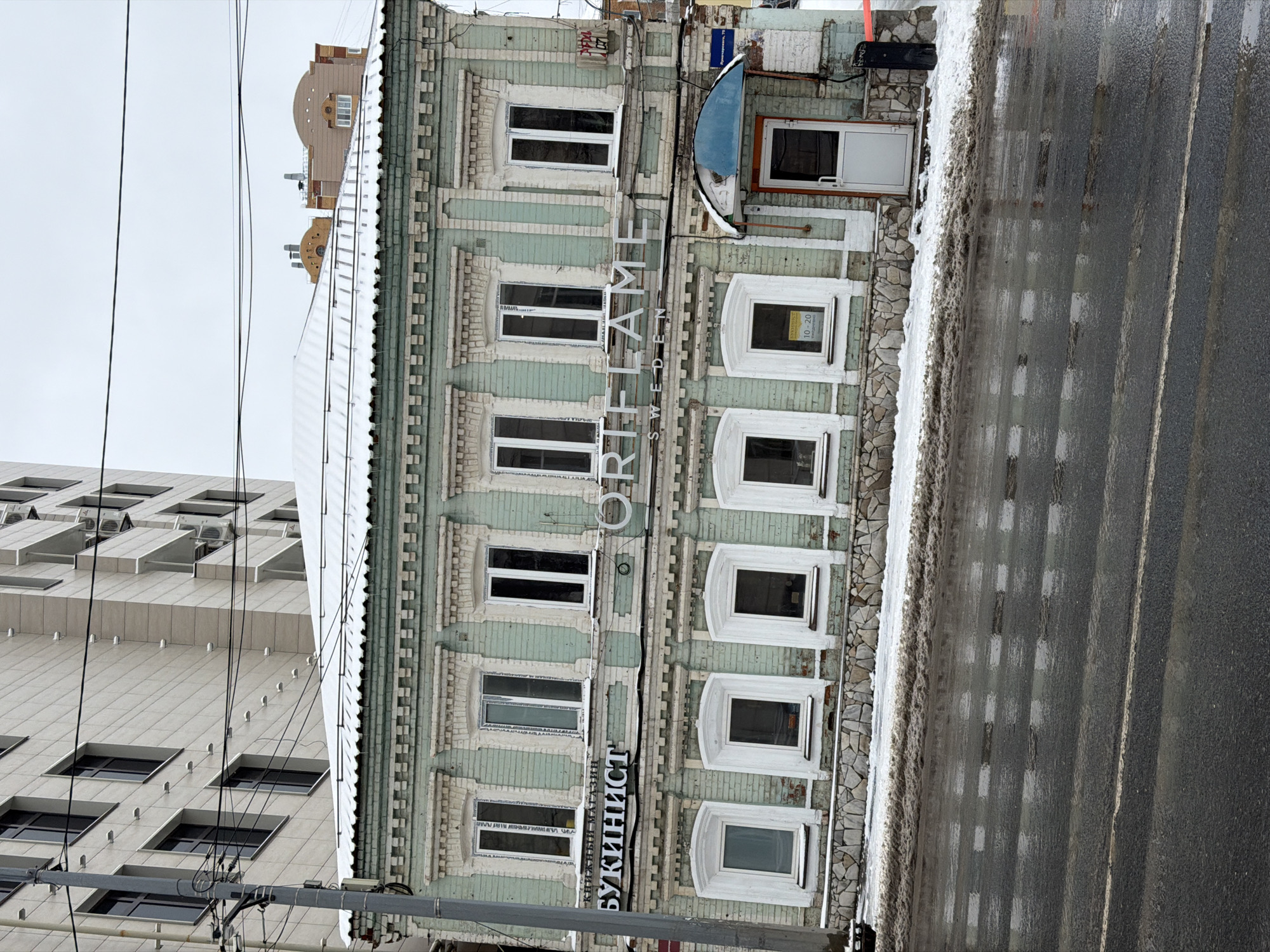 A light-green, four-story building occupies the center of the frame with "Oriflame" vertically emblazoned in white letters along its facade. The sign "Букинист" (Bukinist - Bookstore) is visible at the bottom of the building. A gray sky looms above, while a modern, beige building is seen to the left. The right side of the frame reveals a wet street with some residual snow and slush along the curb. The location is likely the city of Perm, Russia based on the cyrillic sign, the architectural style, and the Oriflame sign.
