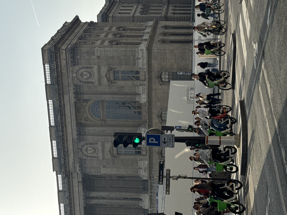 The image depicts a bustling street scene in Paris, France, with the Louvre Museum building prominently featured in the background. The sky is bright and hazy, with a faint aircraft contrail visible in the upper right.
In the background, the large, multi-story Louvre Museum building exhibits classical grey stone architecture, characterized by numerous tall arched and rectangular windows, fluted columns, ornate decorative relief panels with medallions (some bearing a "JL" or "LV" monogram), and a continuous balustrade along its rooftop. At street level in front of the museum, white temporary construction barriers are visible.
In the mid-ground, a dark grey traffic light pole stands to the left of center, displaying an illuminated green light for vehicular traffic. Below it, a blue "P" sign with a white left arrow indicates parking. Further down, white street signs point left to "LOUVRE SAMARITAINE," "GALERIE VERO DODAT," and "QUARTIER JEUNES."
The foreground features an asphalt road with white lane markings and a pedestrian crosswalk. A white bicycle lane symbol is painted on the asphalt to the bottom right. Strong shadows from people and street furniture extend across the road, suggesting a sunny day with light coming from the right.
Numerous individuals populate the lower half of the frame. A crowd of people stands or walks on the left. Several individuals are actively riding electric scooters, predominantly green Lime-branded models, or conventional bicycles. Specifically, from left to right, multiple individuals on Lime scooters, including women in a red top, a light grey hoodie, and a dark top, are moving or waiting to proceed. A woman on a red scooter/moped is also visible. Further right, a bearded man on a dark bicycle is visible. Their attire is casual. Most individuals are facing or moving towards the right side of the frame, indicating movement across an intersection in alignment with the green traffic signal.