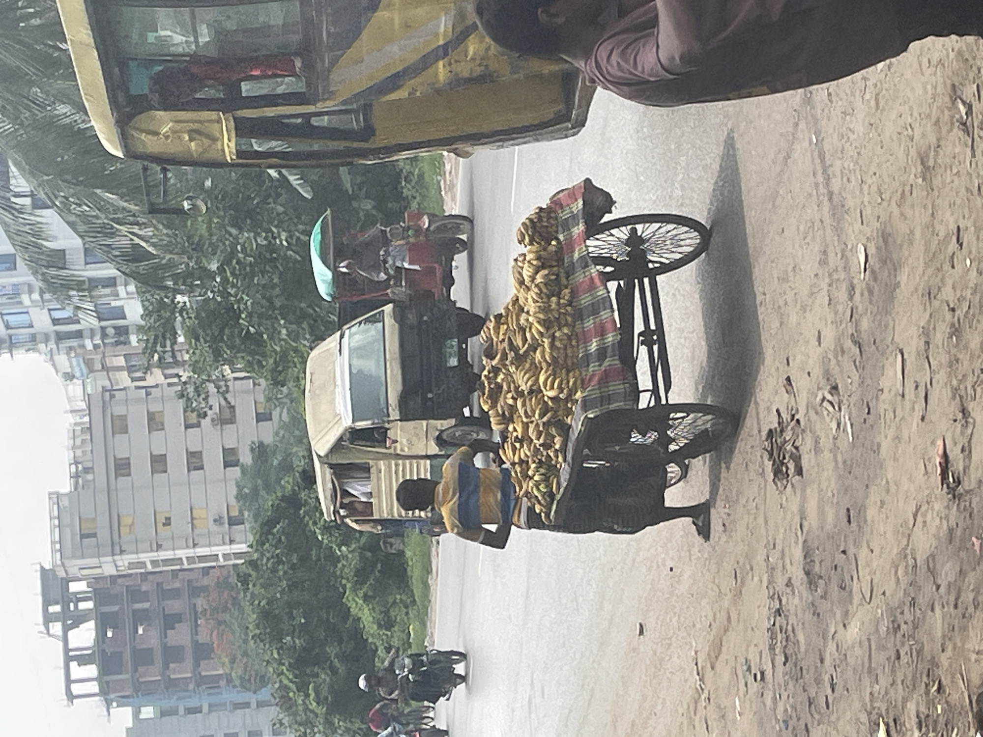 The image depicts a street scene in Dhaka, Bangladesh. A man in a blue and yellow striped shirt is standing next to a three-wheeled cart piled high with bananas, likely a street vendor. A yellow motorized rickshaw, known as a CNG, is partially visible in the foreground. Several motorcycles and other vehicles are visible on the road. Buildings are visible in the background, interspersed with trees. The road appears to be paved, and the overall environment suggests a bustling urban setting.