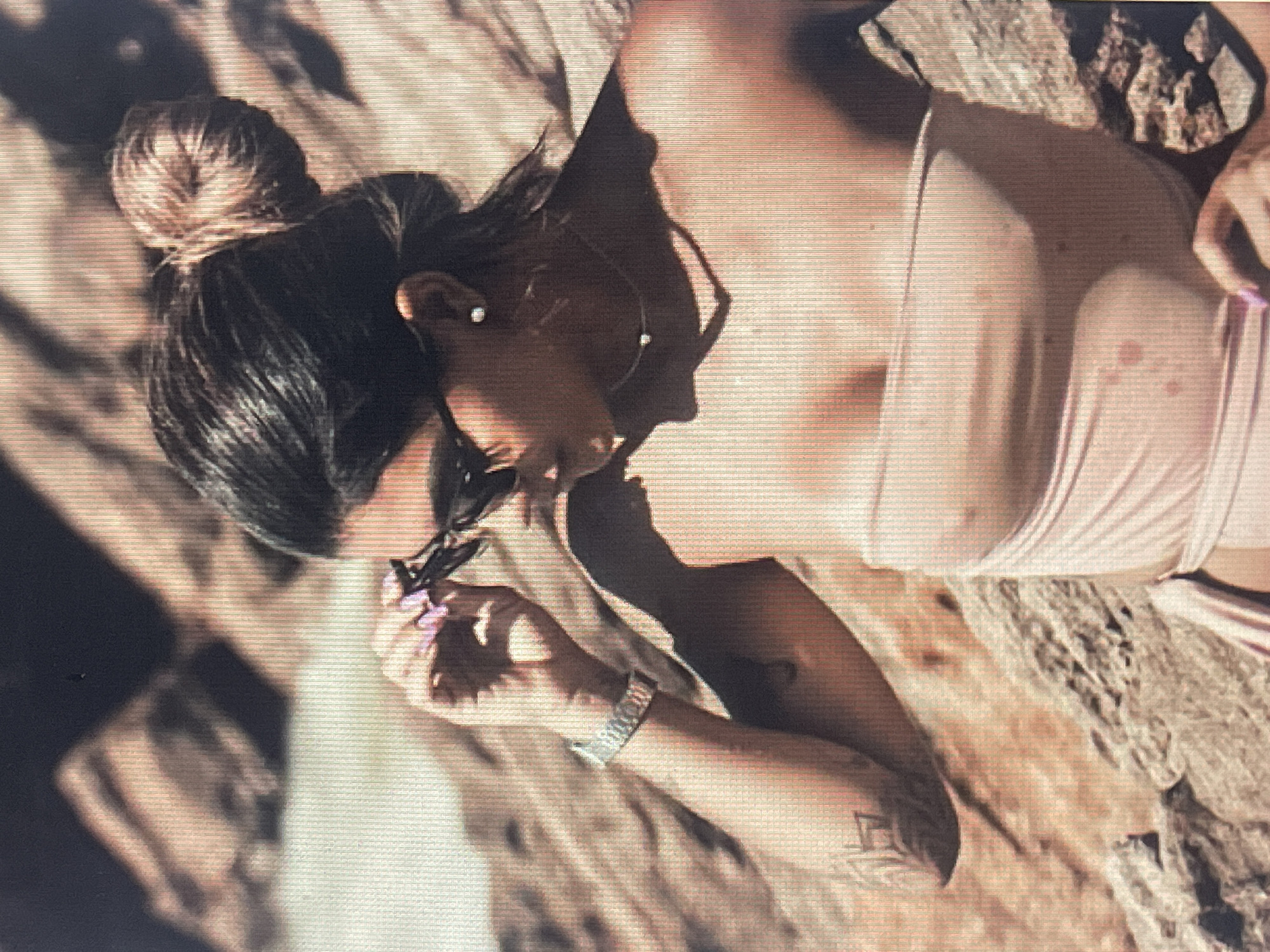 This media file shows a woman lying on rocks, possibly outdoors. The woman has her hair pulled up in a bun and is wearing a strapless top. She is holding a pair of sunglasses and appears to be looking at them. Her nails are painted purple. She is also wearing a watch on her left wrist. The lighting suggests it might be daytime, with shadows indicating sunlight. The background consists of textured rocks. The image is somewhat grainy, with visible horizontal lines.