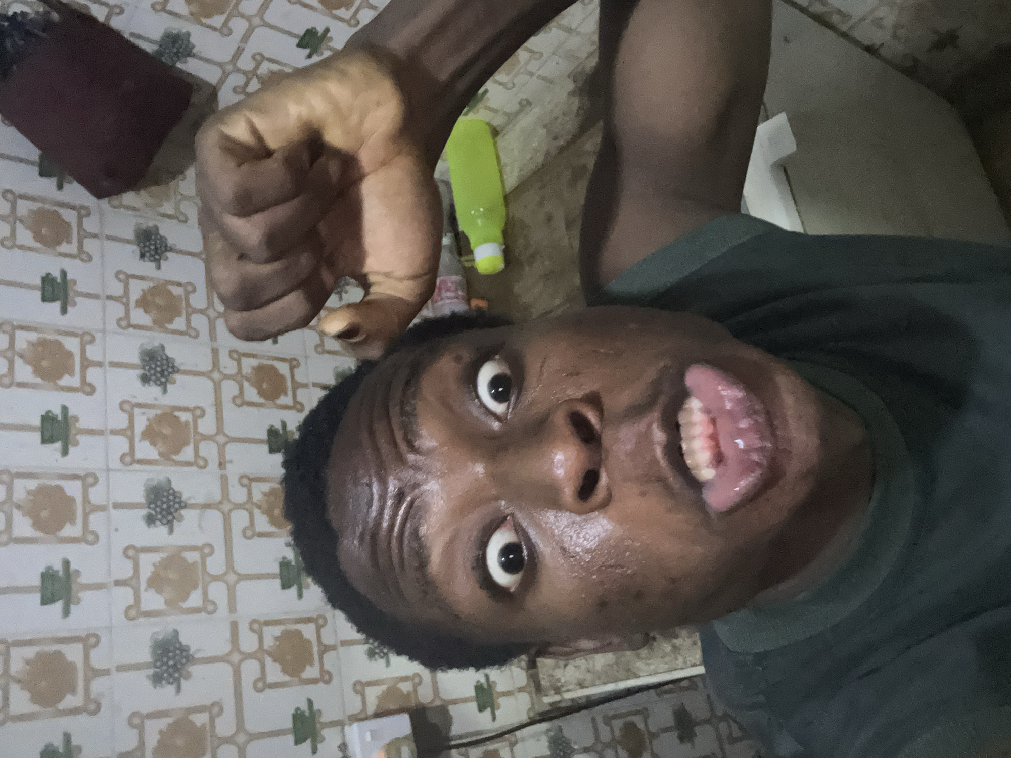 The image is a close-up, selfie-style shot of a person's face and upper body, seemingly taken from a low angle. The person has dark skin, close-cropped black hair, and is wearing a dark-colored shirt. Their eyes are wide open and they are sticking their tongue out slightly, giving a playful expression. Their skin appears shiny, possibly due to sweat. A clenched fist is held up in the top left corner of the frame. The background is a tiled floor with a repeating pattern of beige and green geometric shapes. Scattered on the floor are a brown object, a partially visible green bottle, and other indistinct items. The location is Unknown, Unknown.