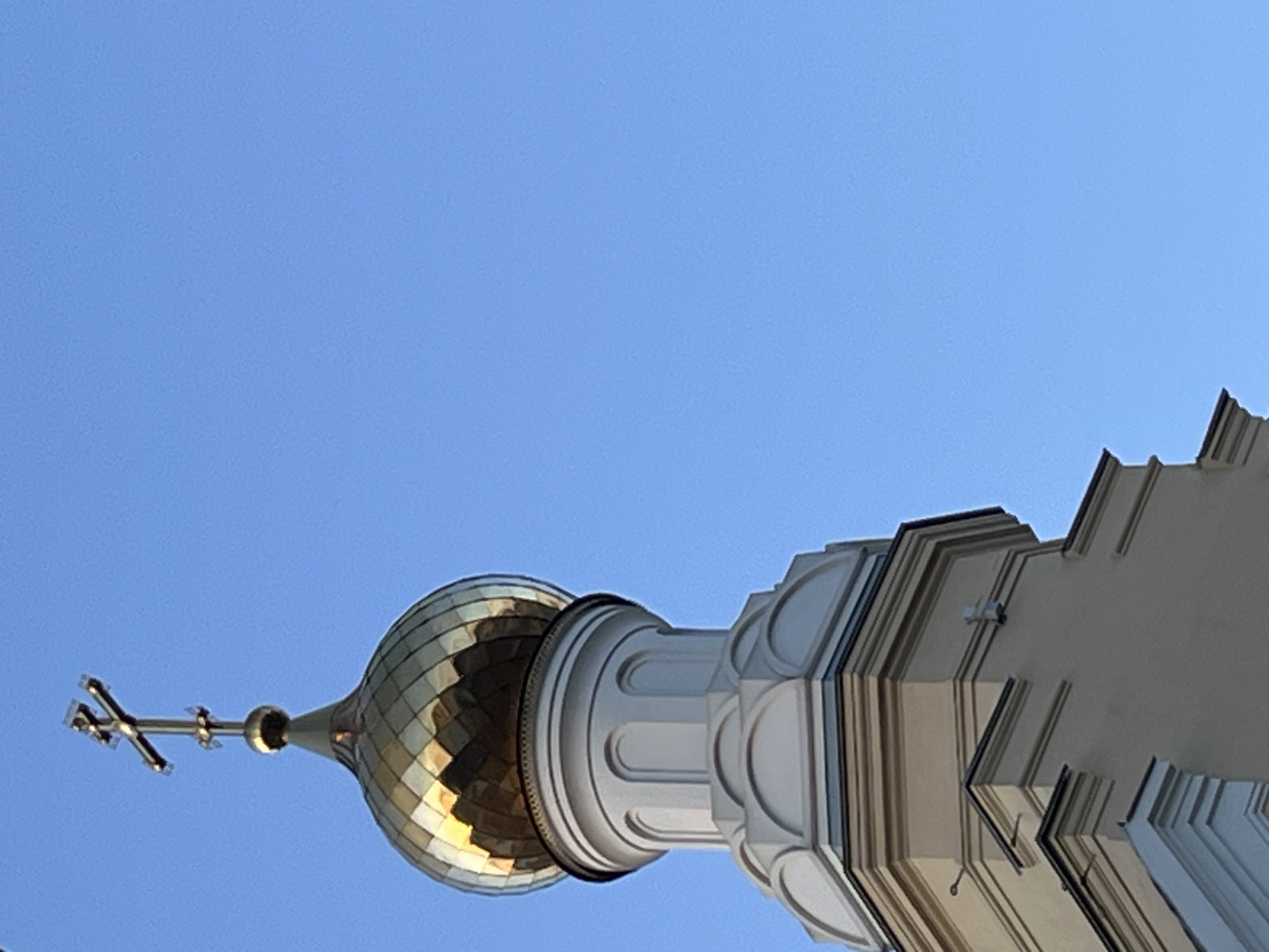 The image is an upward-angled view of the golden dome and cross of an Orthodox church. The dome is golden with a pattern of squares, likely reflecting the sunlight. Atop the dome sits an Orthodox cross with three horizontal bars. To the right of the dome, we see the architectural details of the church building itself, with white or light-colored features and distinct ledges and lines. The background is a clear blue sky. The location is Unknown, Unknown.