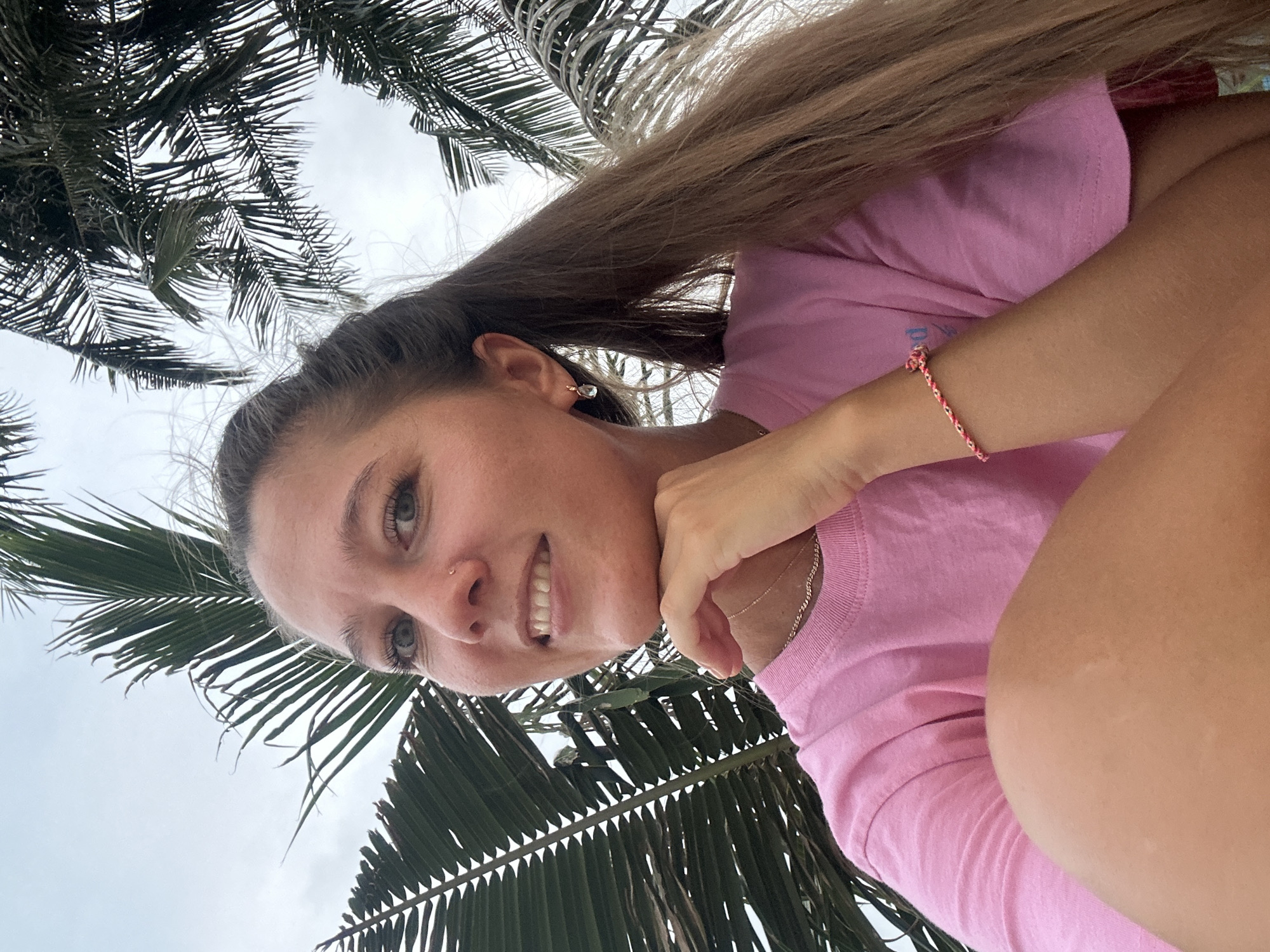 An outdoor, low-angle, medium shot captures a young woman posing for a selfie amidst palm fronds, likely in Ko Samui District, Thailand. She is positioned in the lower-right of the frame, with her face filling the upper-left quadrant. The woman has long, light brown hair pulled back into a ponytail, with strands falling forward around her face. She has fair skin, light blue eyes, a small nose ring, and her left hand is resting under her chin, supporting her face. She is smiling, revealing her teeth, and has a subtle blush on her cheeks. She is wearing a bright pink t-shirt, a delicate gold necklace, and a thin gold hoop earring in her left ear. A braided pink and red bracelet adorns her left wrist. Her right arm is bent, with her knee visible in the lower-left corner of the frame. The background consists of the overcast sky and the dark green, feathery fronds of palm trees, suggesting a tropical or subtropical location. The lighting is soft and diffused, consistent with an overcast day.