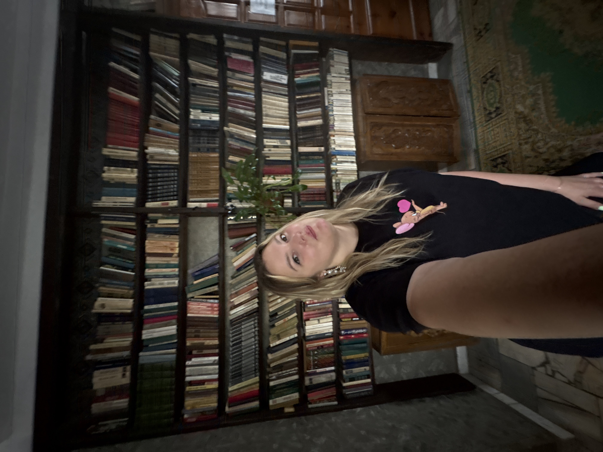 A woman with blonde hair and large earrings is taking a selfie in front of a large, dark wood bookshelf filled with books. She is wearing a black t-shirt with a cartoon character and hearts printed on it. The bookshelf is tall and spans the width of the wall, with multiple shelves packed with books of various sizes and colors. In the foreground, a portion of the woman's arm and torso are visible. Below the bookshelf, there are two intricately carved wooden cabinets. A patterned rug is visible on the floor in front of the cabinets. The overall impression is an interior scene, likely a home or library, with a focus on books and the woman's presence. The image is captured from a slightly low angle, looking up at the woman and the bookshelf. There is no visible indication that this scene is located in Zavod Mikhailovskii, Russia.