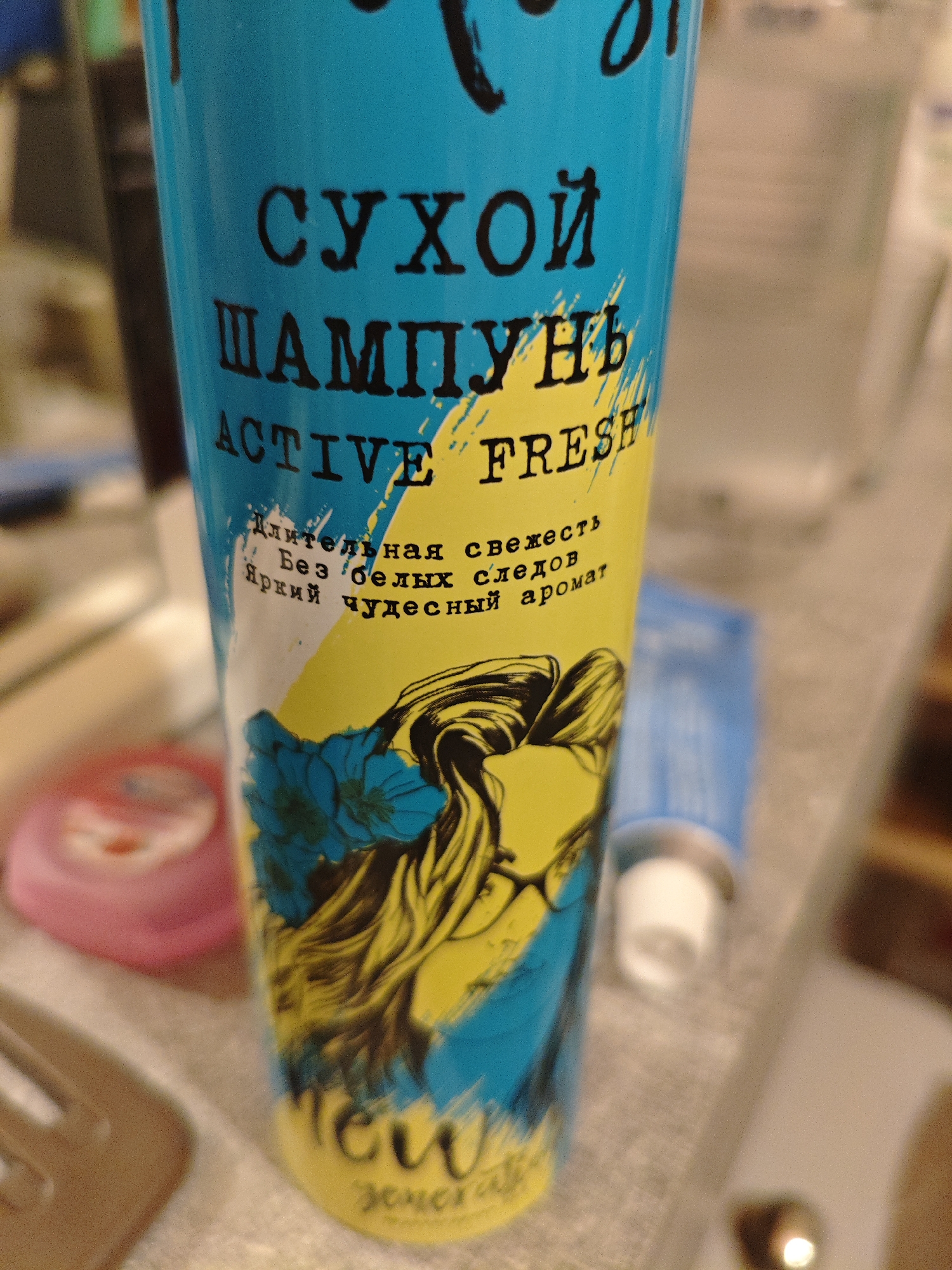 The image shows a close-up of a blue and yellow aerosol can, likely a dry shampoo product, placed on a surface. The can features prominent text in Russian and English. The Russian text reads "СУХОЙ ШАМПУНЬ," which translates to "DRY SHAMPOO." Below this, the English text "ACTIVE FRESH" is visible, along with smaller Russian text that says "Длительная свежесть. Без белых следов. Яркий чудесный аромат," meaning "Long-lasting freshness. No white residue. Bright wonderful aroma." A stylized illustration of a woman's head with long, wavy hair adorned with blue flowers is printed on the can. At the bottom of the can, there appears to be a logo with stylized text, possibly the brand name, with "NEW" in large letters above it. The can is positioned at an angle, with the background blurred and out of focus, showing hints of other objects on what appears to be a countertop or shelf. The lighting suggests an indoor setting, possibly a bathroom or a dressing area. The overall impression is a product shot focusing on the design and key features of the aerosol can. The location context provided is Berdsk municipality, Russia.