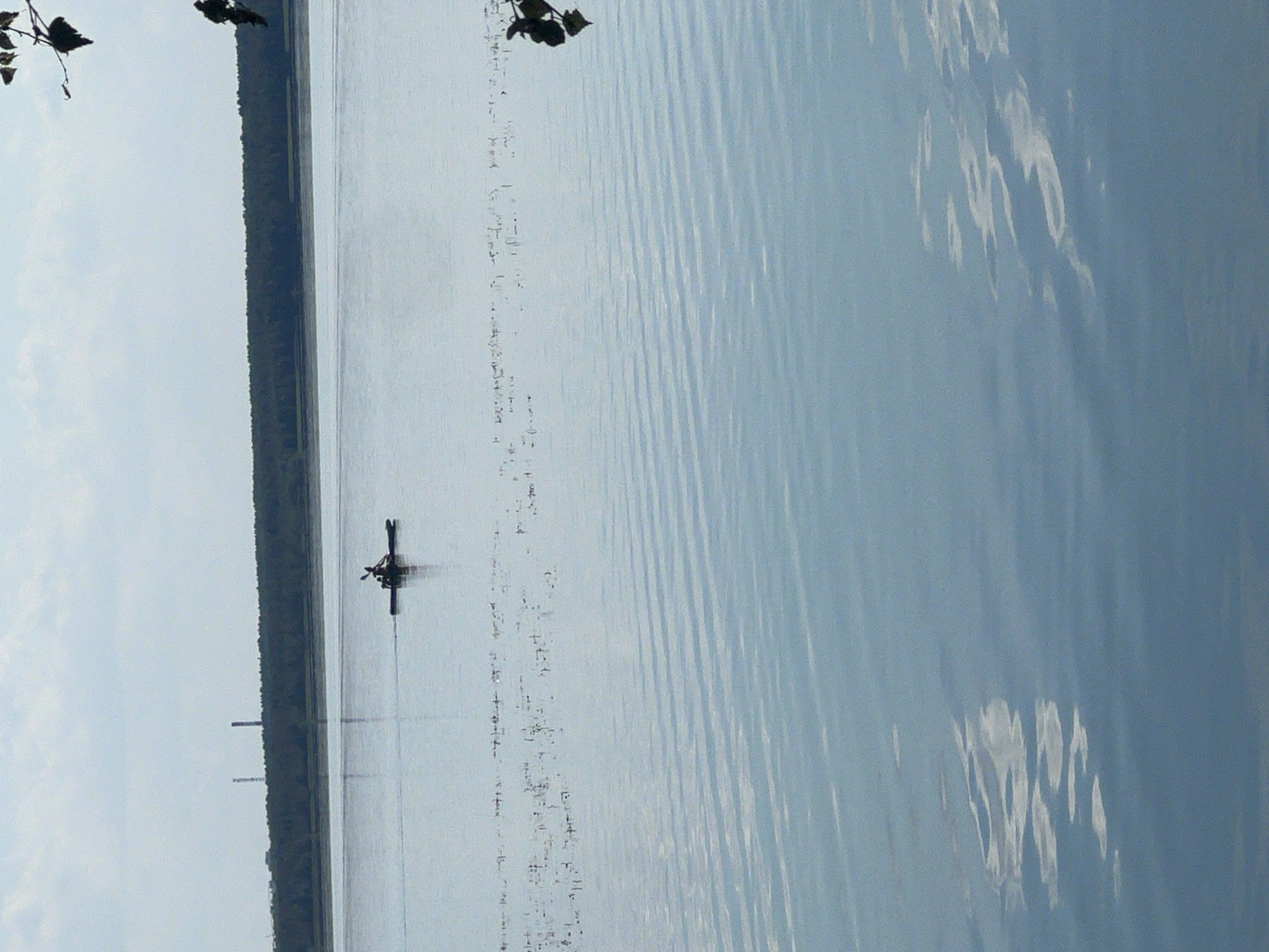 A calm, wide river reflects a pale blue sky with scattered clouds. A dark green forest lines the distant horizon, interrupted by two tall, thin industrial chimneys. In the middle distance, a person is kayaking on the water. The foreground of the image is filled with the rippling surface of the river, with subtle reflections of the sky and clouds. The scene is serene and quiet.