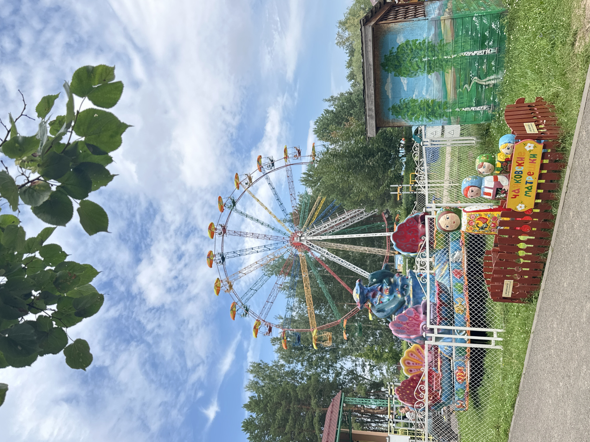 The image depicts a small amusement park in the city of Chaikovsky, Russia. In the background, a colorful Ferris wheel stands tall against a partly cloudy blue sky. In the foreground, a small carousel ride, decorated with aquatic-themed imagery and bright colors, is visible behind a white metal fence. To the right, a small wooden structure is painted with a landscape scene of birch trees and a path. Several matryoshka doll figures are arranged in a wooden enclosure in front of the painted structure. The ground is covered with green grass and a paved pathway is visible in the lower left corner. A few people are present, with one person visible on the carousel and another near a building on the left. The overall scene suggests a recreational area on a sunny day.