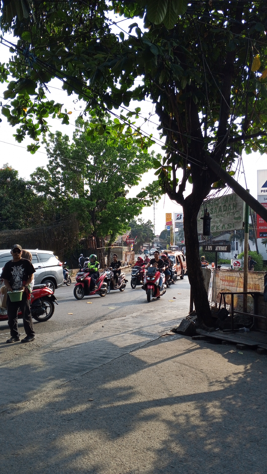 The image depicts a street scene in Bekasi, Indonesia, likely during daylight hours given the bright sunlight casting long shadows. A group of people on motorcycles are traveling along a paved road. One individual, wearing a black t-shirt and a cap, stands on the left side of the road, holding a green bucket. Several motorcycles are in motion, including a red one in the foreground with a rider wearing a dark shirt and a passenger in the back. Another red motorcycle further down the road has a rider with a green helmet and a dark jacket. Behind them, more motorcycles and a van are visible, creating a moderate flow of traffic. The roadside is lined with trees, some with dense green foliage, and electrical wires are strung overhead. Buildings and signs are present in the background, suggesting an urban or suburban environment. One sign appears to be in Indonesian, mentioning "Jatirawat" which is a common street name in Bekasi. The ground is paved and shows signs of wear and tear, with some cracks and patches. The overall atmosphere suggests a typical day on the road in a bustling Indonesian city.