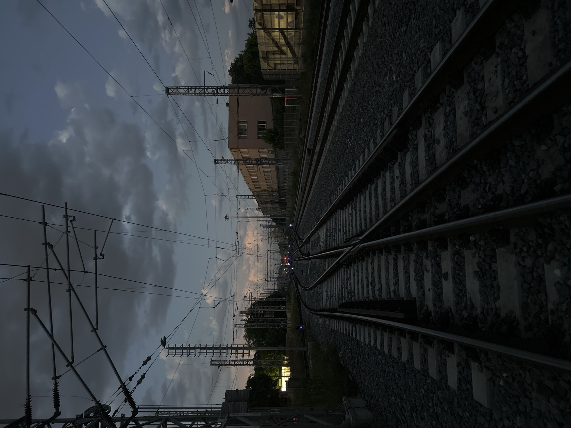 The image depicts a railway scene at dusk. Multiple sets of train tracks converge in the distance, with overhead electrical lines and support structures visible above. To the left of the tracks, a multi-story building is present, potentially an industrial or administrative structure. Vegetation is interspersed along the sides of the tracks. In the distance, faint red and white lights are observable. The image is dark, and it is difficult to definitively identify the specific location in Plovdiv without further information. However, based on the presence of electrical lines and rail infrastructure, it is likely an industrial or transportation zone within the city.