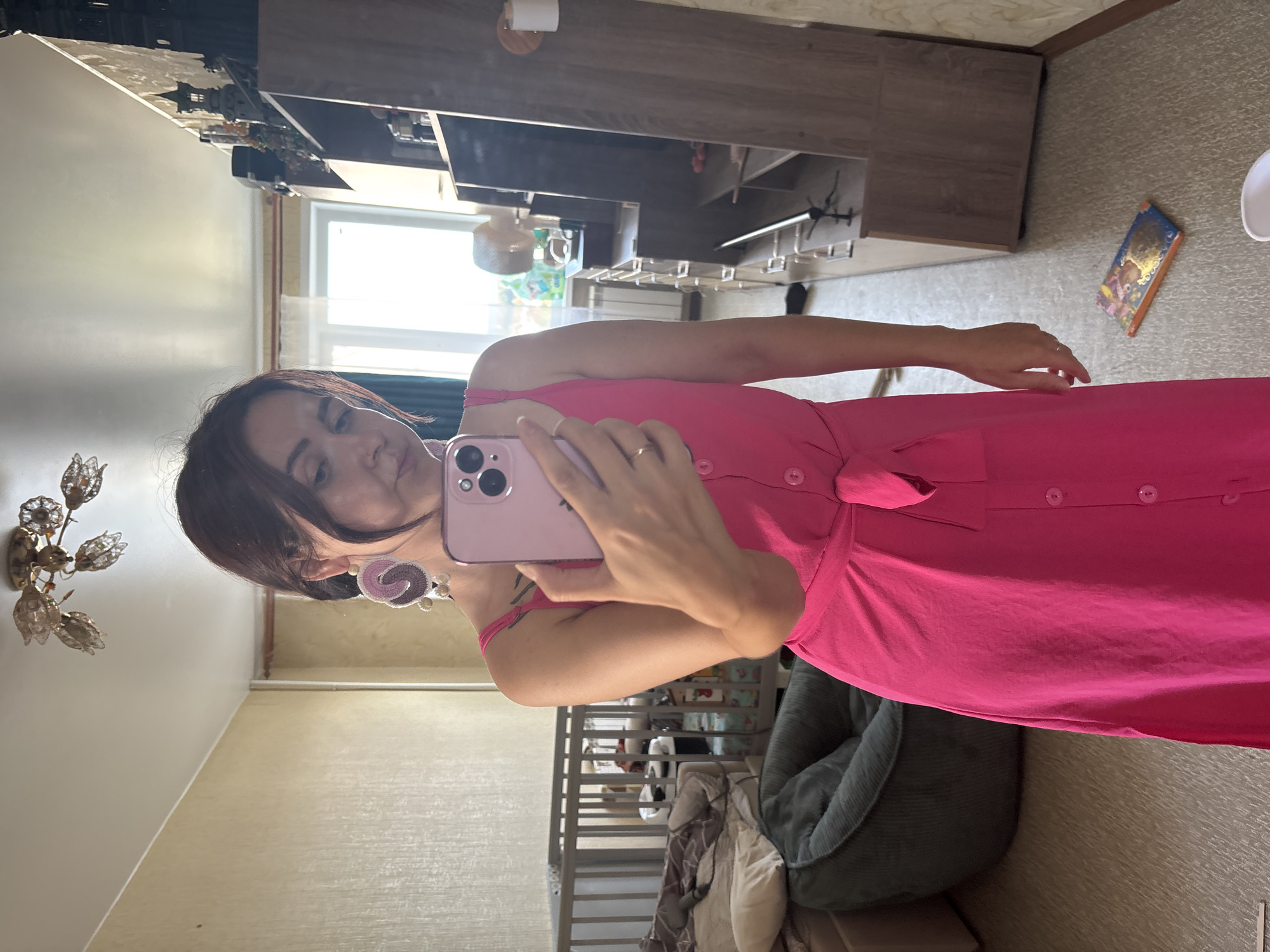 A woman is taking a mirror selfie in a room. She is wearing a bright pink dress with a tie waist and buttons down the front. She has large, circular, crocheted earrings in shades of pink and brown. She is holding a pink smartphone in her right hand and her left hand is resting on her hip. In the background, there is a crib with bedding, a green beanbag chair, a wooden dresser with shelves filled with various items including decorative castle figures, and a window with sheer curtains. The room appears to be a bedroom or nursery. The overall setting suggests a domestic environment, likely a home. It is not possible to determine the specific location in the city of Naberezhnye Chelny, Russia from the visual information provided in the image.