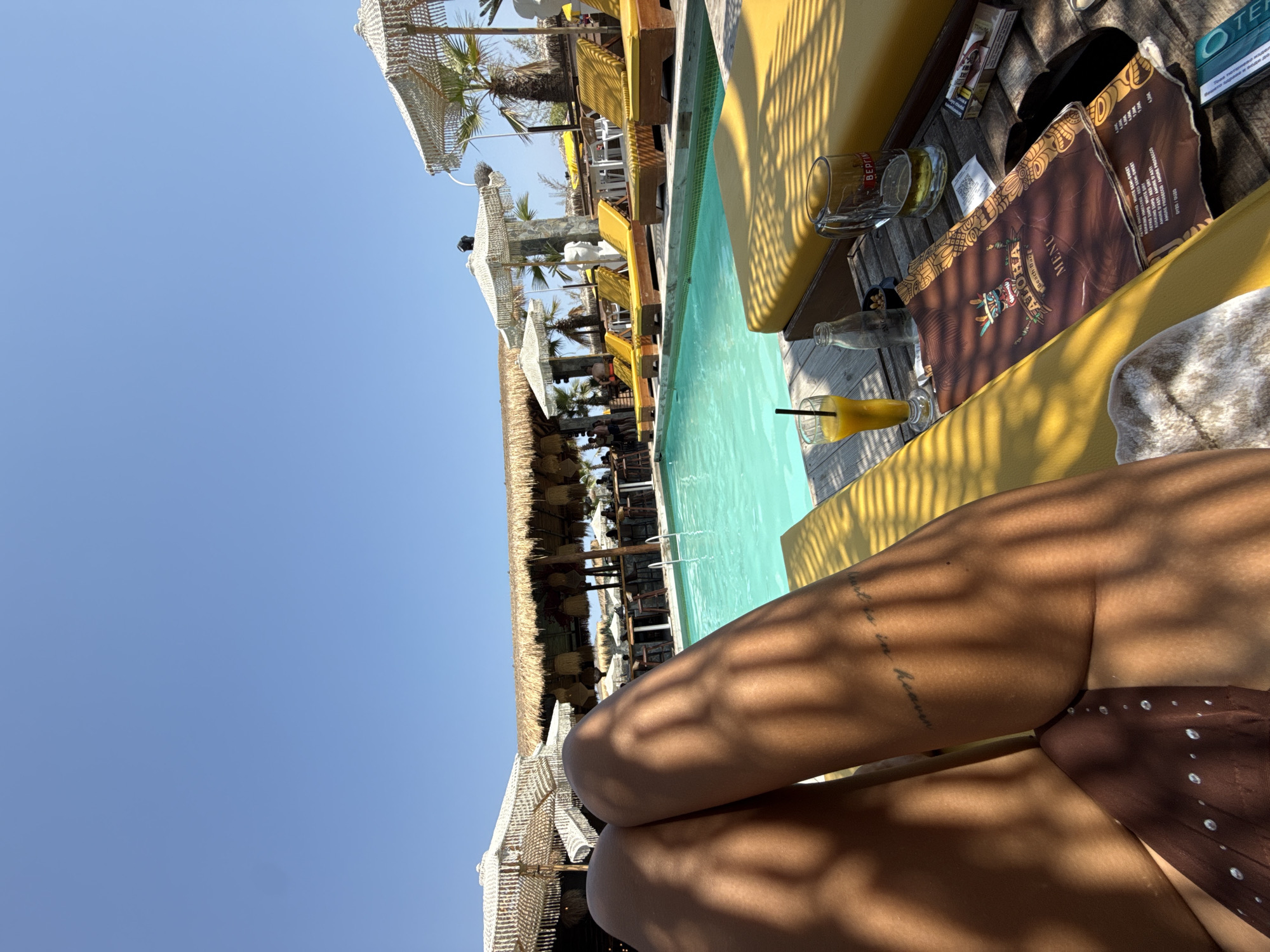 The image depicts a sun-drenched scene at a beach bar or resort in Katerini Municipality, Greece, likely during daytime given the clear blue sky and bright sunlight. The foreground is dominated by the tanned legs of a person reclining on a yellow lounge chair, with a subtle tattoo visible on their left thigh. To the right of the person's legs, a wooden table holds several items. These include a glass of orange juice with a straw, a glass bottle, a beer mug containing a light-colored liquid, a cigarette pack, and a menu for "ALOHA BEACH BAR". The menu's cover features a tribal-style illustration.
Beyond the table, a rectangular swimming pool with turquoise water is prominent, reflecting the bright sky. The pool area is lined with more yellow lounge chairs, many occupied or ready for use. Several thatched-roof umbrellas and palm trees are visible, contributing to a tropical ambiance. In the background, a bar structure with a thatched roof and hanging lamps can be seen, with patrons and staff present. The overall atmosphere is one of leisure and relaxation in a seaside resort setting.