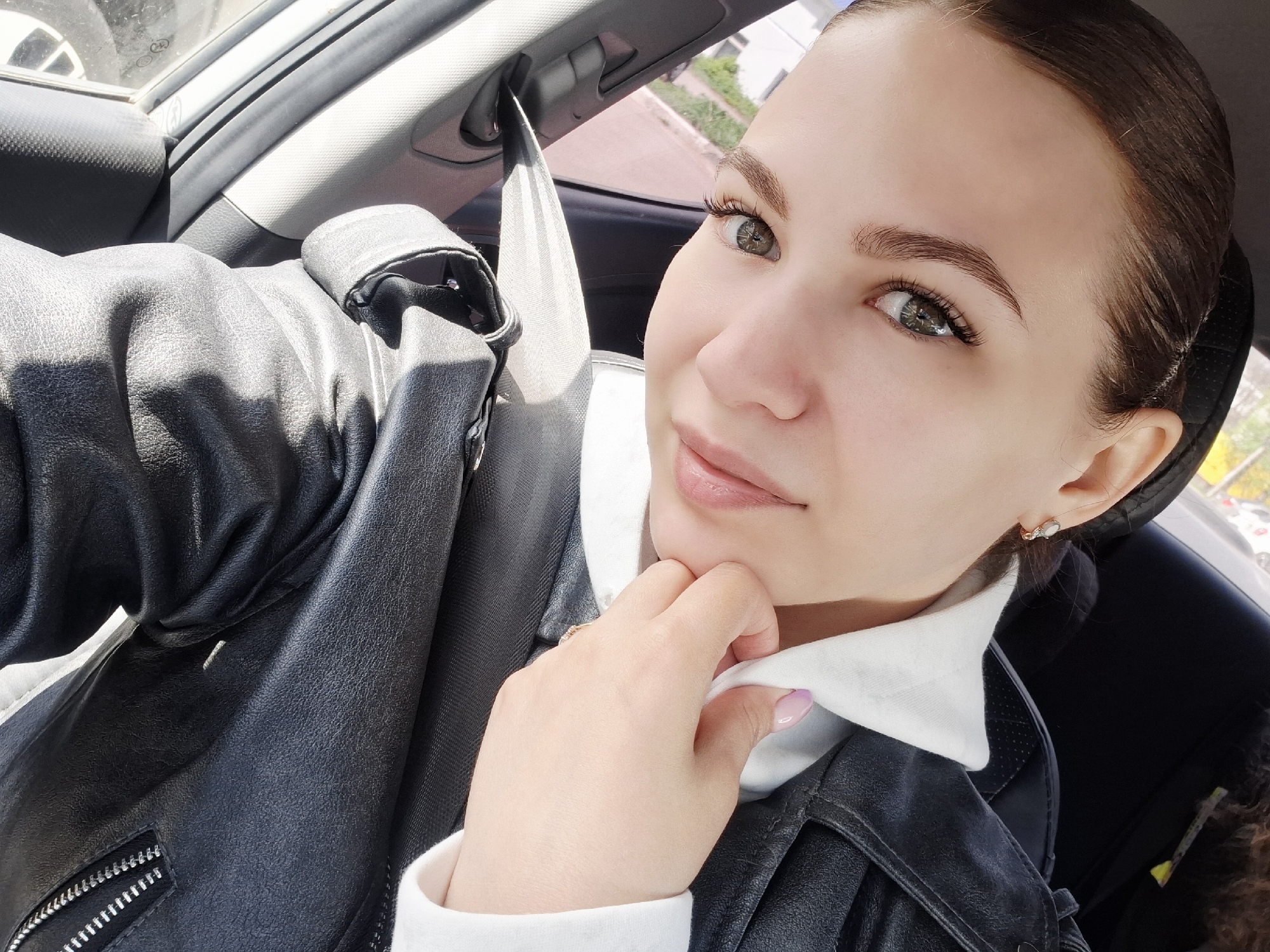 A young woman with brown hair pulled back and wearing a black leather jacket over a white collared shirt is sitting in a car. She is wearing a seatbelt. Her hand is gently touching her chin. The interior of the car is visible around her. The location is Chaikovsky, Russia.