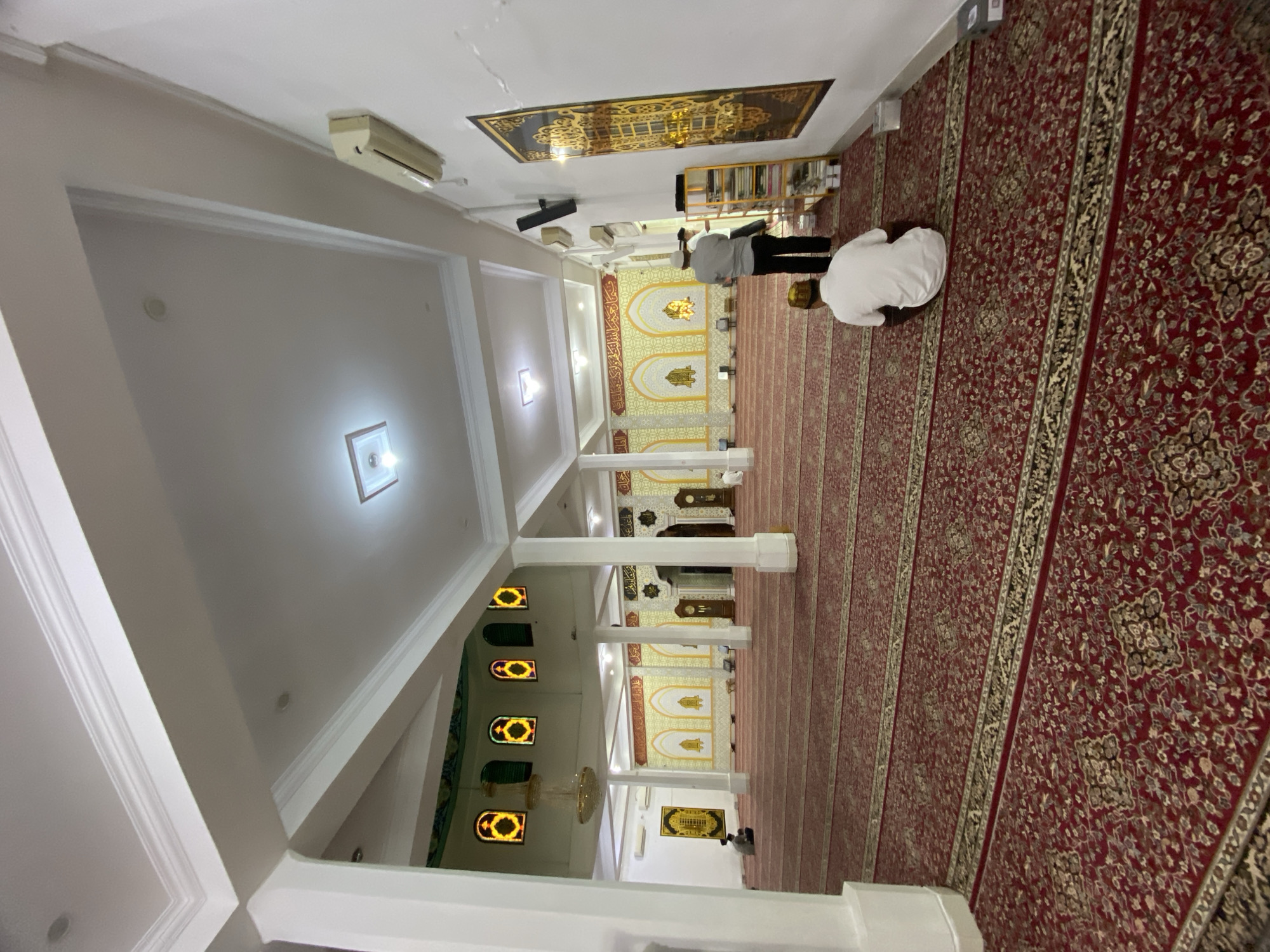 The image shows the interior of a mosque in Lhokseumawe, Indonesia. The mosque has a large, open prayer hall with a red carpet covering the floor. The carpet has a repeating pattern of intricate floral designs. White columns support a white ceiling with recessed lighting. Stained-glass windows with yellow and orange patterns are visible in the distance. Two men are present in the prayer hall. One man is kneeling on the carpet in the foreground, facing away from the camera. Another man, wearing a white shirt and black pants, stands near a shelving unit on the right side of the image. A large, ornate tapestry hangs on the wall to the right. The overall impression is one of a clean and well-maintained place of worship.
