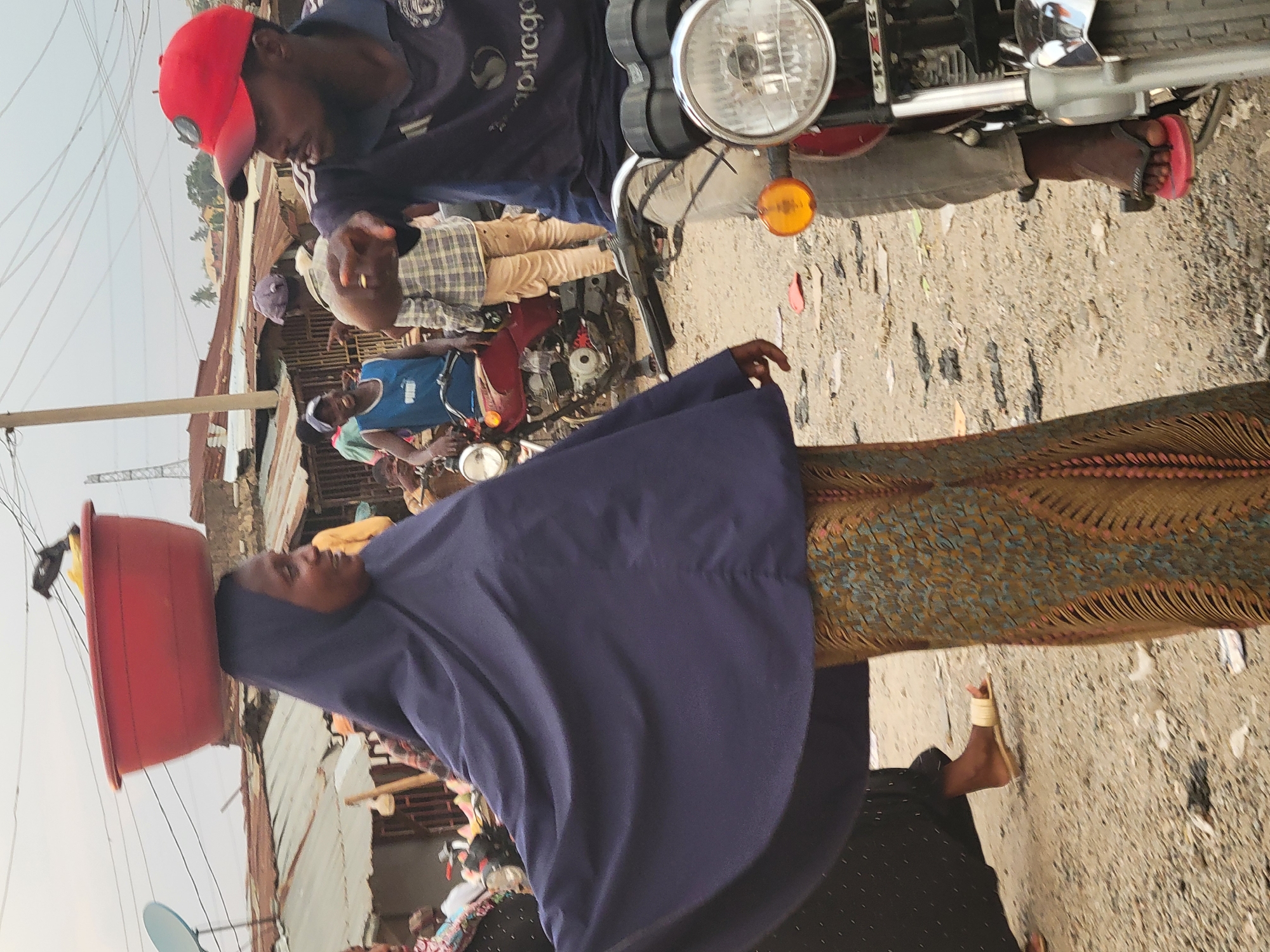 In Madalla, Nigeria, the scene depicts a woman carrying a red basin on her head, wearing a navy blue hijab and a patterned brown and blue dress. A man in a red cap and blue shirt stands near her, close to a motorcycle with its headlight visible. Additional people are present in the background. The environment appears to be a busy street or market area.