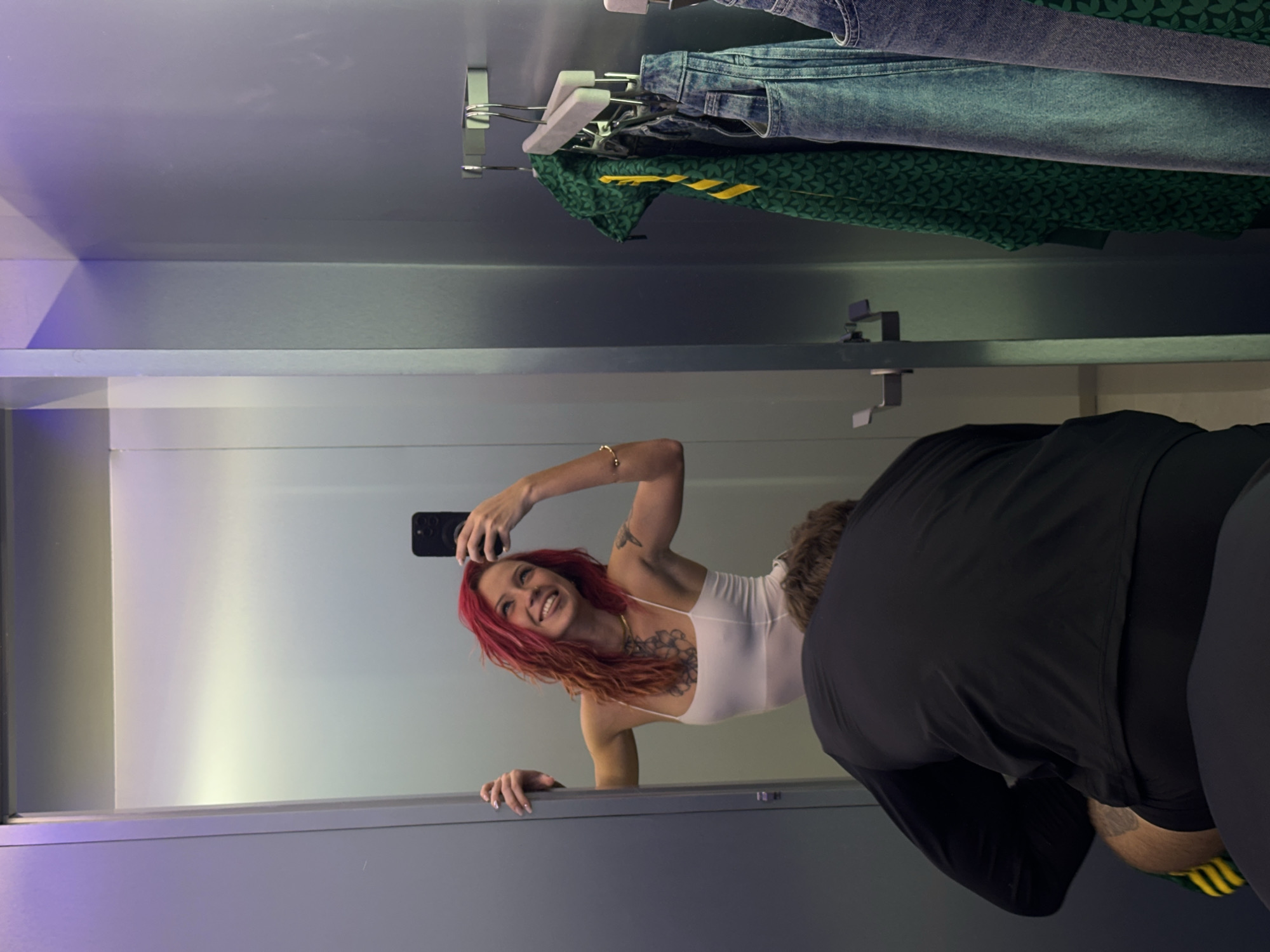 In a store fitting room, a woman with red hair is reflected in a mirror, taking a selfie with a smartphone. She is wearing a white tank top and has tattoos on her upper body. To the right of the mirror, a person wearing black athletic clothing is bent over, partially obscuring the view. Above the mirror, clothing, including jeans and a green jacket, hangs on a rack. The fitting room has a gray and white color scheme. There is no information in the image that supports the claim the scene is in Kuta, Indonesia.