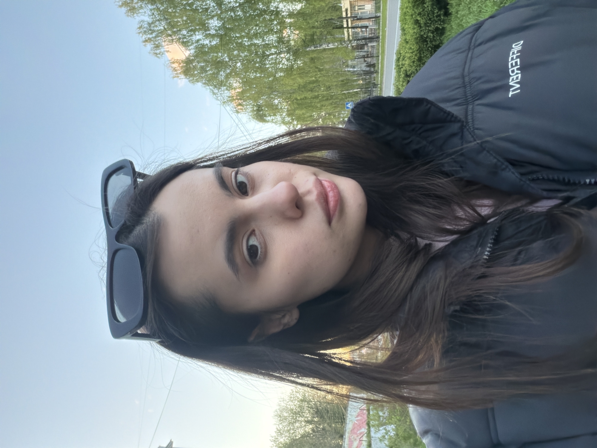 A young woman is taking a selfie outdoors. She has long brown hair and dark sunglasses perched on her head. She is wearing a dark puffer jacket. The background suggests an urban environment, with trees, buildings, and a road visible. Based on the available information, it's not possible to confirm if the image was taken in the city of Chaikovsky, Russia or any other specific location.