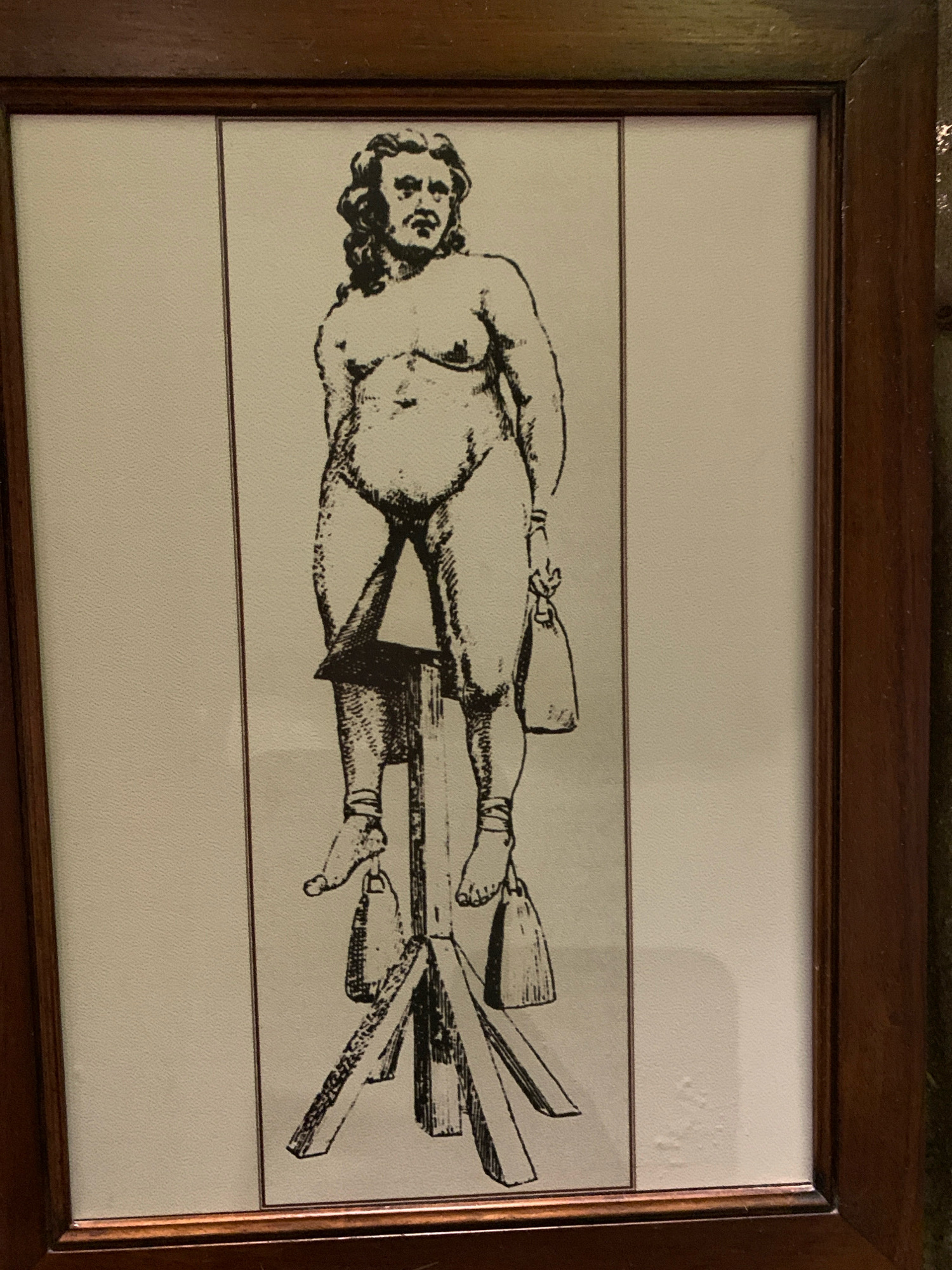 The image is a black and white illustration framed in wood. The illustration depicts a nude male figure standing with his feet elevated on a wooden structure. Bags are suspended from his ankles. The figure has long, wavy hair and a mustache. The background is a plain, light-colored surface with a subtle textured pattern. The overall scene appears to be an artistic representation of a historical or possibly symbolic subject. There is no discernible information within the image or its framing that indicates its location in the City of San Marino, San Marino.