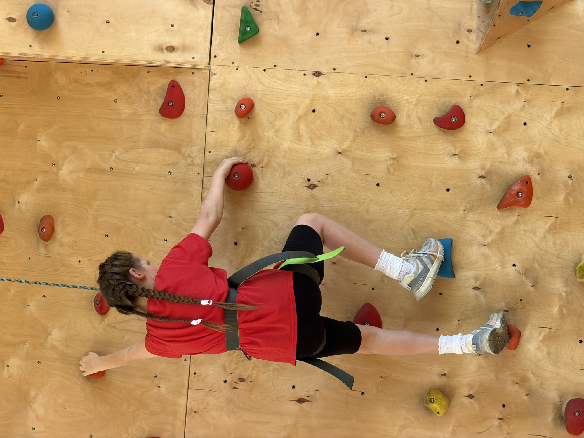 A young person is indoor rock climbing on a plywood wall, wearing a red shirt, black shorts, climbing shoes, and a harness with a green loop. The wall has numerous colored climbing holds attached to it. There is a climbing rope visible. The location is an indoor climbing wall, likely in Izhevsk, Russia.