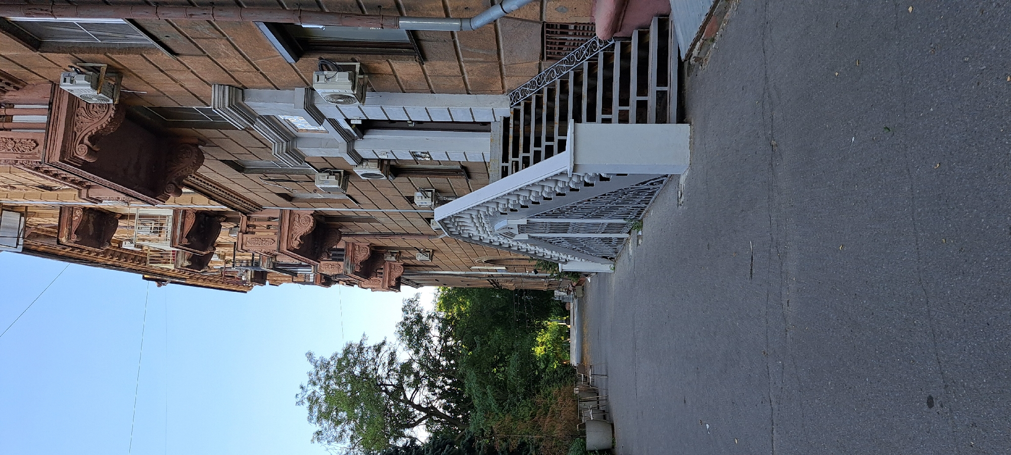 The image shows a side view of a brick building with ornate architectural details. There are several air conditioning units mounted on the exterior walls. A white staircase with intricate metal railings leads up to an entrance. To the right of the building, there is an asphalt walkway or road. In the background, lush green trees and a clear blue sky are visible. The lighting suggests it is daytime, with sunlight casting shadows. There are no people or animals visible in the image. The scene appears to be an exterior shot of a residential or commercial building in an urban or suburban setting. The focus is on the building's facade and the architectural elements.