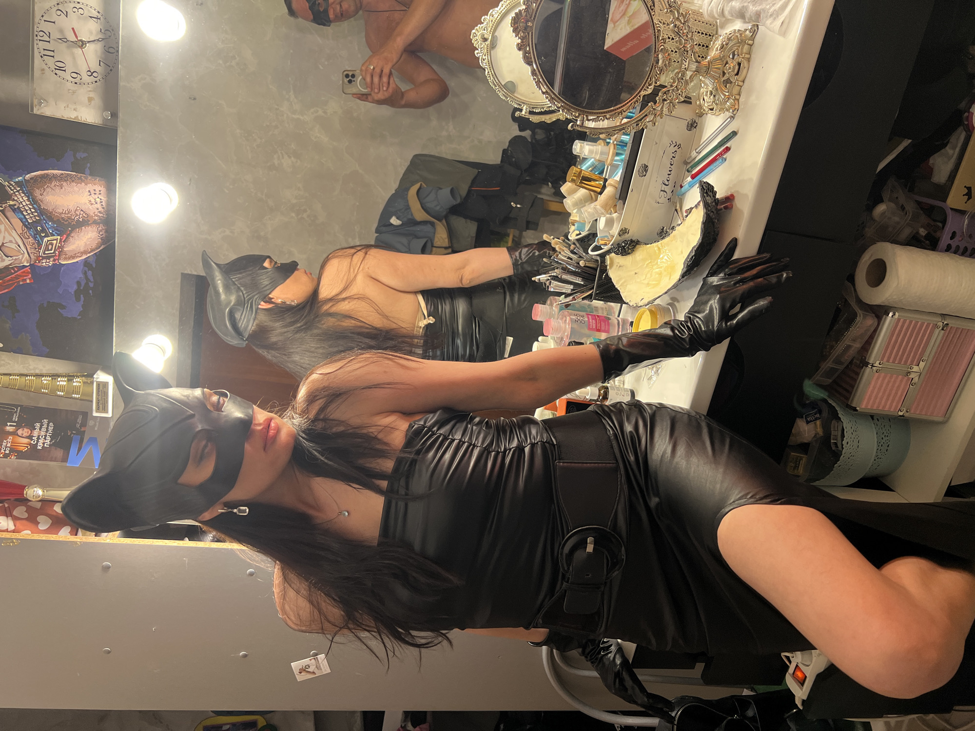 A woman in a black cat-ear mask and a black strapless leather dress is posing in a room. She is wearing black gloves and a thick black belt. The room appears to be a dressing room or vanity area, with a mirror reflecting another person taking a photo with a phone. There are various makeup and beauty products on a counter, including brushes, bottles, and a decorative mirror. In the background, there are framed pictures and a clock. The presence of a "Flowers" labeled container and a pink metal case suggests a personal or professional grooming space, potentially backstage at an event. The woman's posture and the overall scene convey a sense of playful theatricality, possibly related to a costume or performance. The location in Perm, Russia, is indicated by signage in the background, including Russian text on a framed item reading "УСПЕХ ВО ВСЁМ" (Success in Everything) and "САМЫЙ КРАСИВЫЙ ПАРТНЕР" (Most Beautiful Partner).
