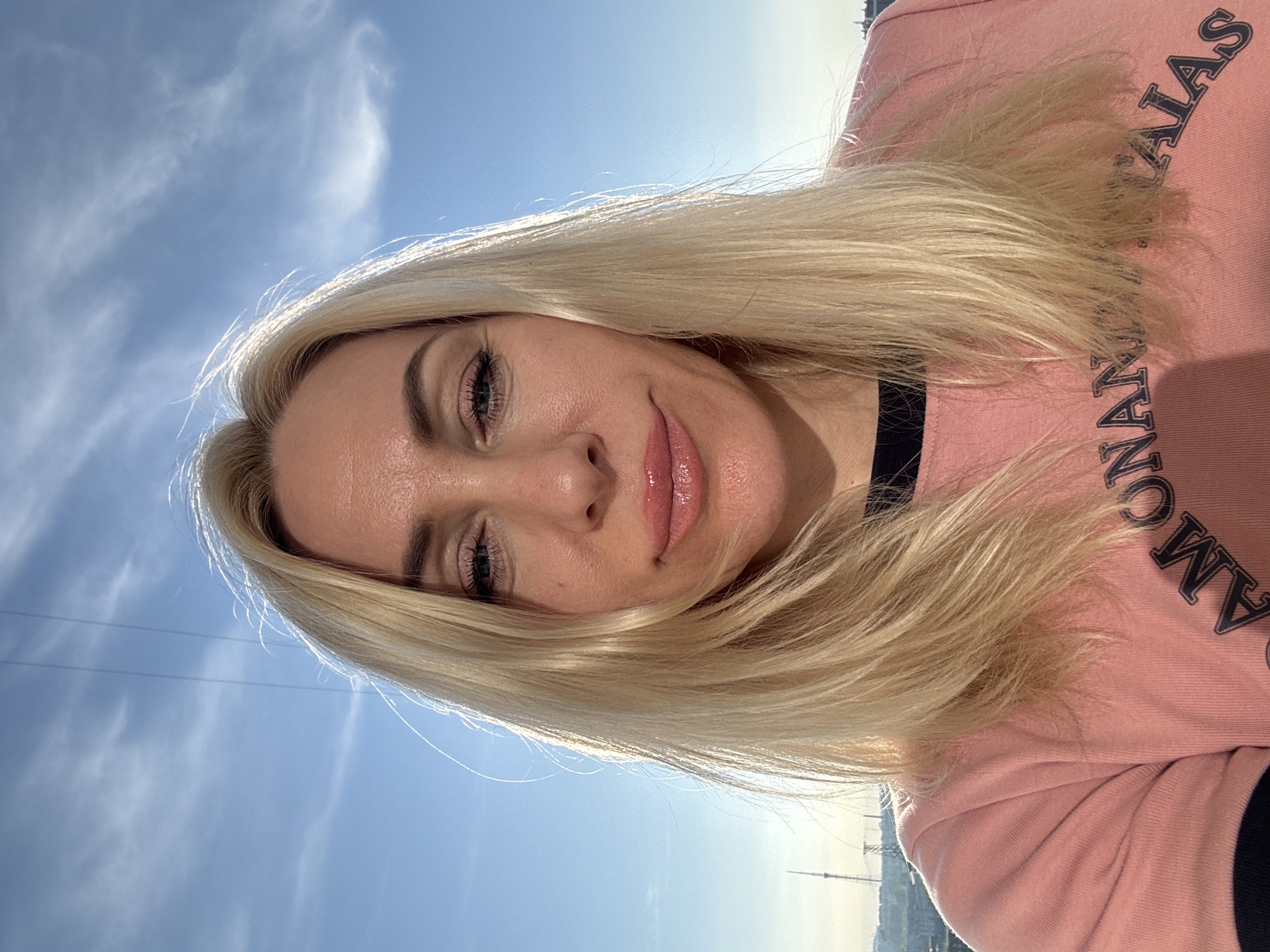 A low-angle shot features a fair-skinned woman with blonde hair, wearing a pink shirt with black lettering. The sky in the background is light blue with clouds. The woman's expression is a slight smile. Part of a cityscape is visible in the distant background.