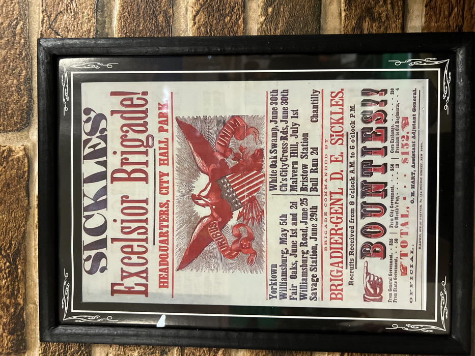 A framed vintage poster advertises Sickles' Excelsior Brigade, headquartered at City Hall Park. It lists various battles, including Yorktown, Williamsburg, Fair Oaks, and Bull Run 2nd. The brigade is commanded by Brigadier-General D.E. Sickles. Recruits were received from 8 a.m. to 6 p.m. The poster prominently displays "Bounties," with monetary amounts listed from general, state, and city governments, along with corn exchange and premium to old regiments, totaling $152. The official is O.H. Hart, Assistant Adjutant General, with printing by Baldwin, Stein Printer, NYC.