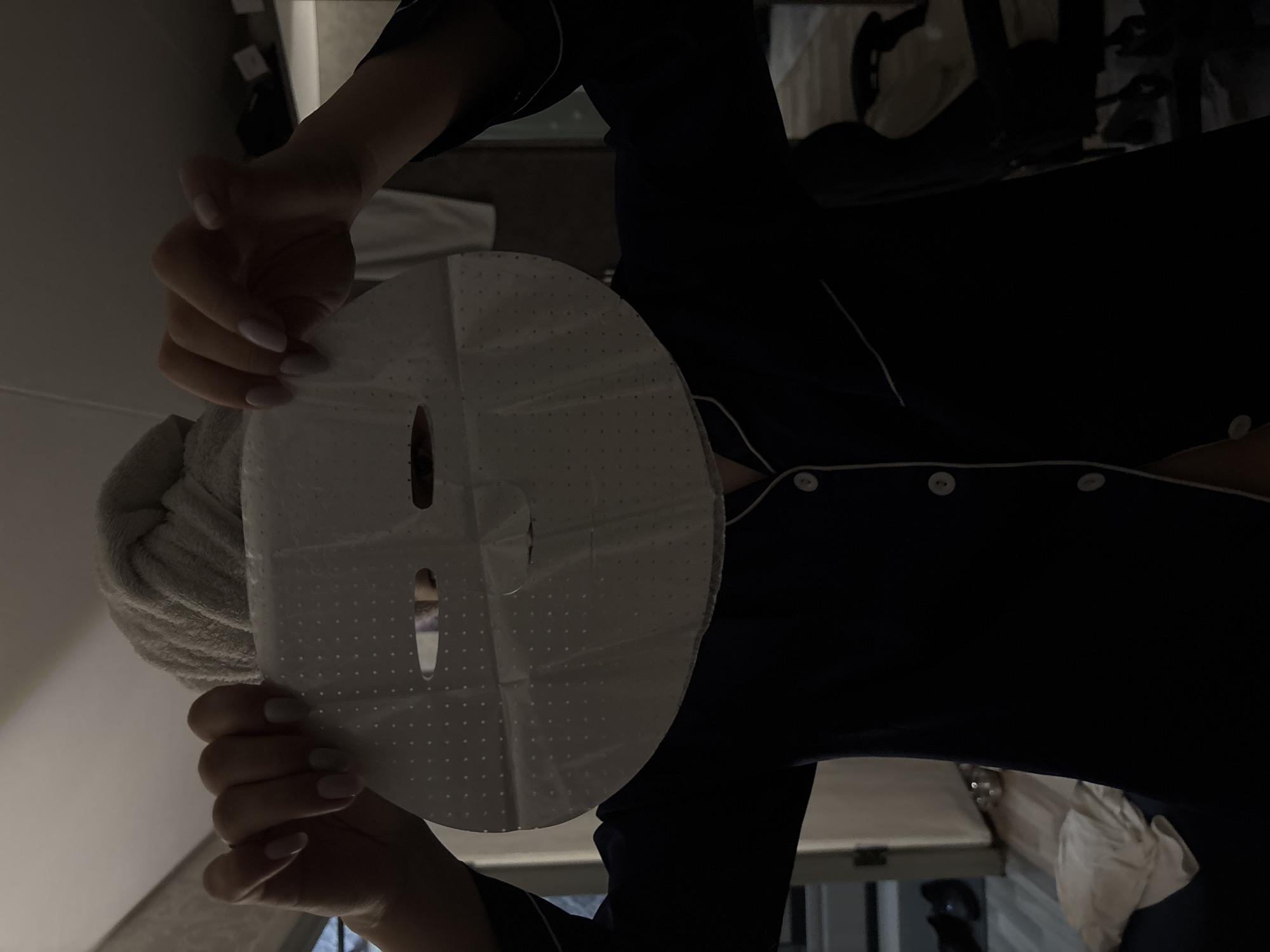 The image captures a person in an indoor setting, engaged in a self-care routine. The subject is holding a white sheet face mask directly in front of their face, obscuring facial features. Both hands are visible, supporting the mask. The mask itself is white with a pattern of small, embossed dots and includes cutouts for the eyes, nose, and mouth.
The person is wearing dark-colored pajamas with light-colored piping detail along the seams and white buttons down the front. A light-colored, possibly beige or grey, towel is wrapped around their head, indicating recent hair washing or preparation for a beauty treatment.
The scene is dimly lit, suggesting it is evening or nighttime. The background is dark and out of focus, providing no specific details about the room's decor or furnishings, but confirms an enclosed, private space. No text is visible anywhere in the image. The overall impression is one of personal care and relaxation. The provided location context is Chaikovsky, Russia.