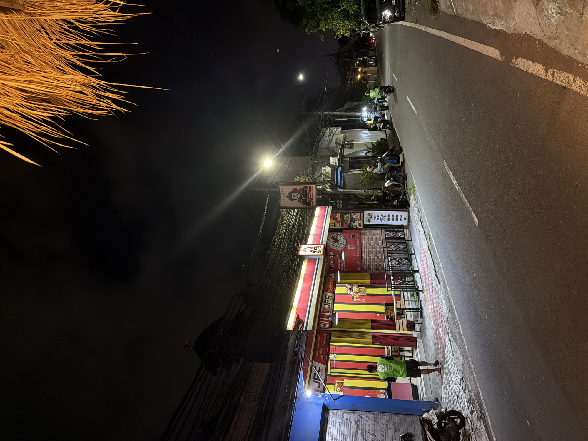 At night in North Kuta, Indonesia, a JFS restaurant with red and yellow striped walls is open for business. A person wearing a green Gojek uniform stands outside the restaurant. Scooters are parked along the sidewalk and on the road. Streetlights illuminate the road and nearby buildings. The road has a white dividing line. Trees and wires are visible above the restaurant and on the left side of the frame.