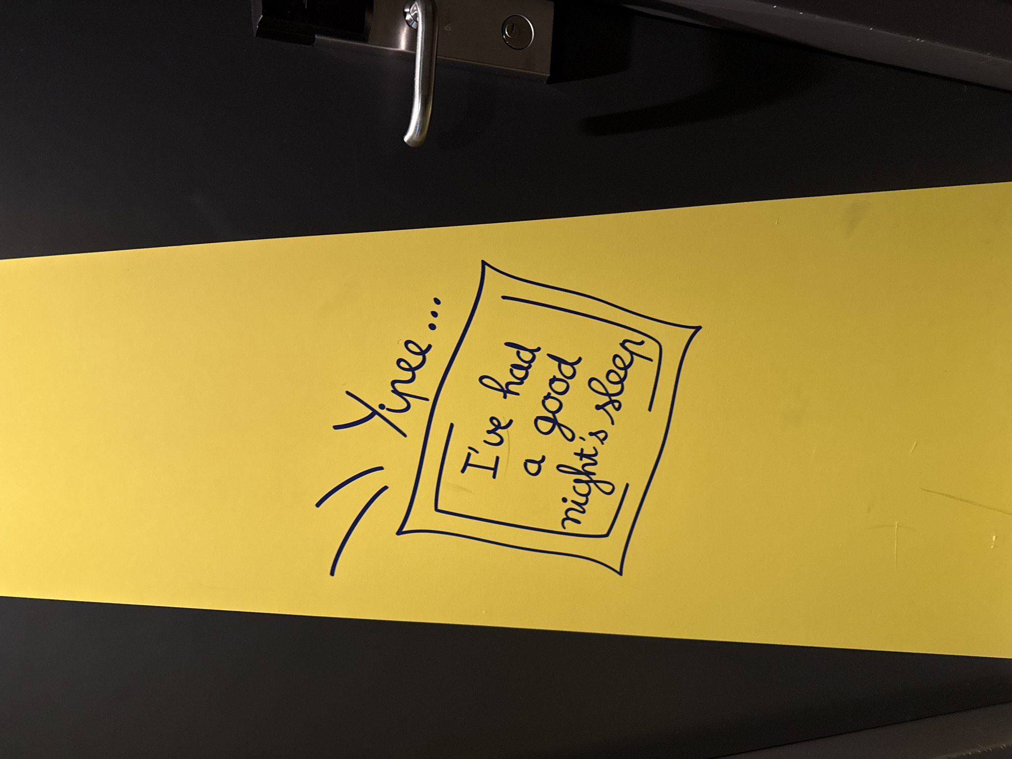 The image displays a yellow vertical panel with dark blue handwritten text. At the top, the text "Yipee..." is written, followed by a box containing the message "I've had a good night's sleep". The yellow panel is positioned next to a dark grey surface, which appears to be a door. A silver door handle and a lock mechanism are visible on the dark grey surface. The overall scene is indoors, suggesting it may be a hotel room or similar accommodation. No people are present in the image.
