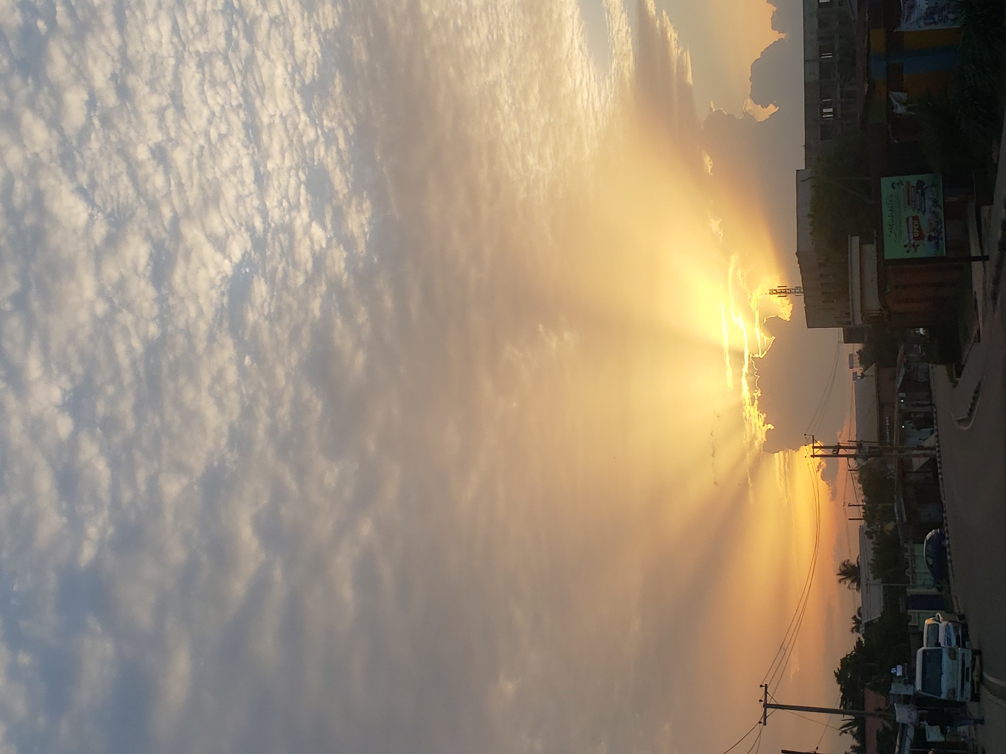 A view from a street in Narayi, Nigeria, shows a dramatic sunset with sunbeams radiating from behind clouds. The sky is filled with scattered, textured clouds, transitioning from a pale blue and grey at the top to a warm golden orange near the horizon. The sun itself is obscured by clouds, but its light creates a powerful visual effect.
In the foreground, a paved road curves to the right, bordered by a sidewalk. On the left side of the street, a light-colored van is parked, with a person walking away from it. Further down the street, other vehicles are visible, including a dark blue sedan. Utility poles with electrical wires stretch across the scene.
The background features a collection of buildings, some appearing to be under construction with exposed frameworks, while others are more complete. Vegetation, including trees and bushes, is present throughout the scene, softening the urban landscape. A billboard with colorful graphics and text, including the word "OPEN," is visible on the right side of the street. The overall impression is of a busy street scene during a beautiful sunset.